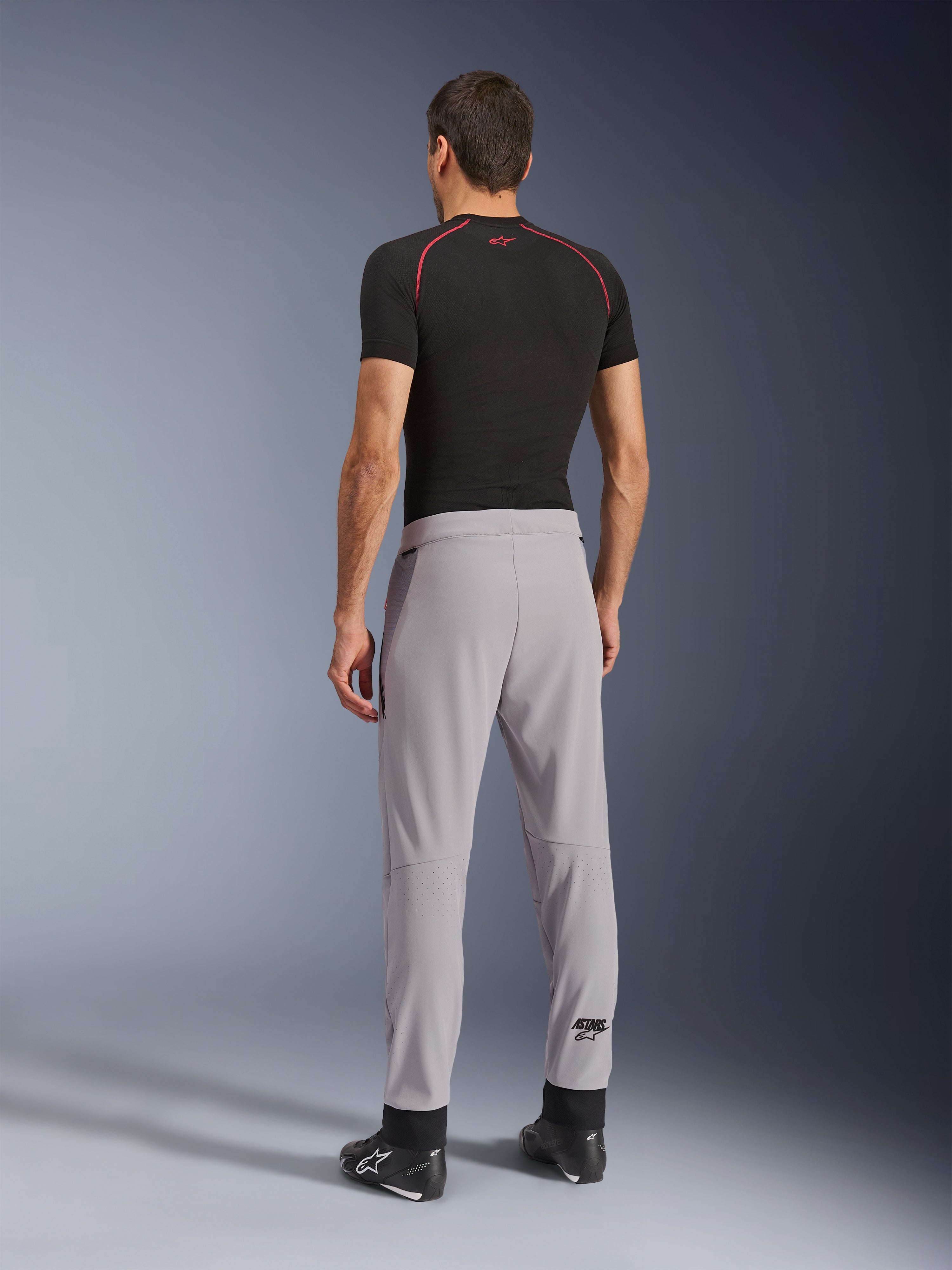 Bakifrån av en modell som bär Alpinestars Aeroshell Airflow-byxor i Mid Gray, kombinerat med en svart underställströja och svarta skor, vilket framhäver de laserperforerade zonerna på vaderna och den atletiska sportpassformen.