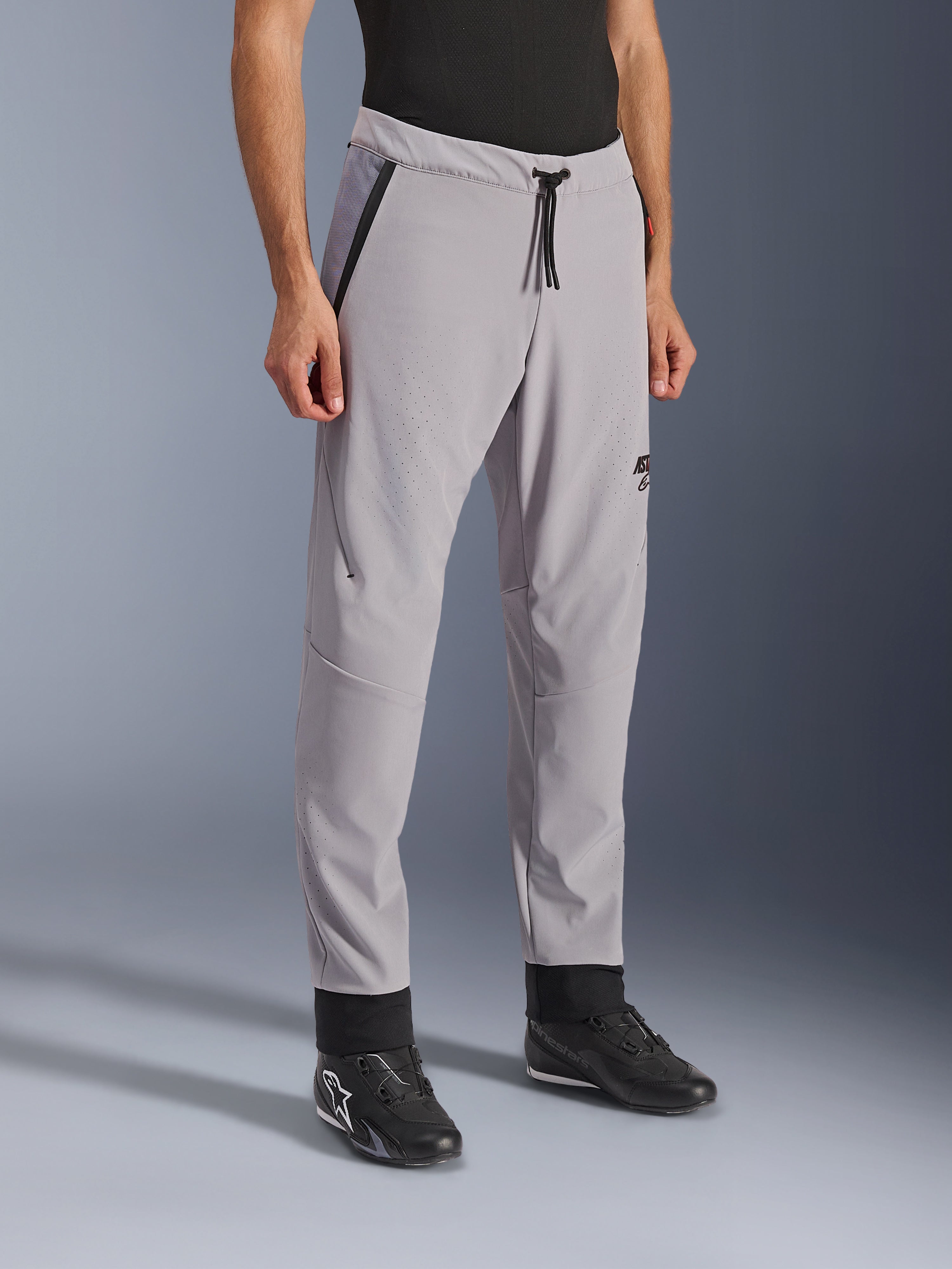 Alpinestars Aeroshell Airflow Pants, Mid Gray, lätta motorcykelbyxor med laserperforerade ventilationszoner på lår och smalben, elastiskt midjeband med dragsko och smal sportig passform, visas på en modell som bär svarta körskor.