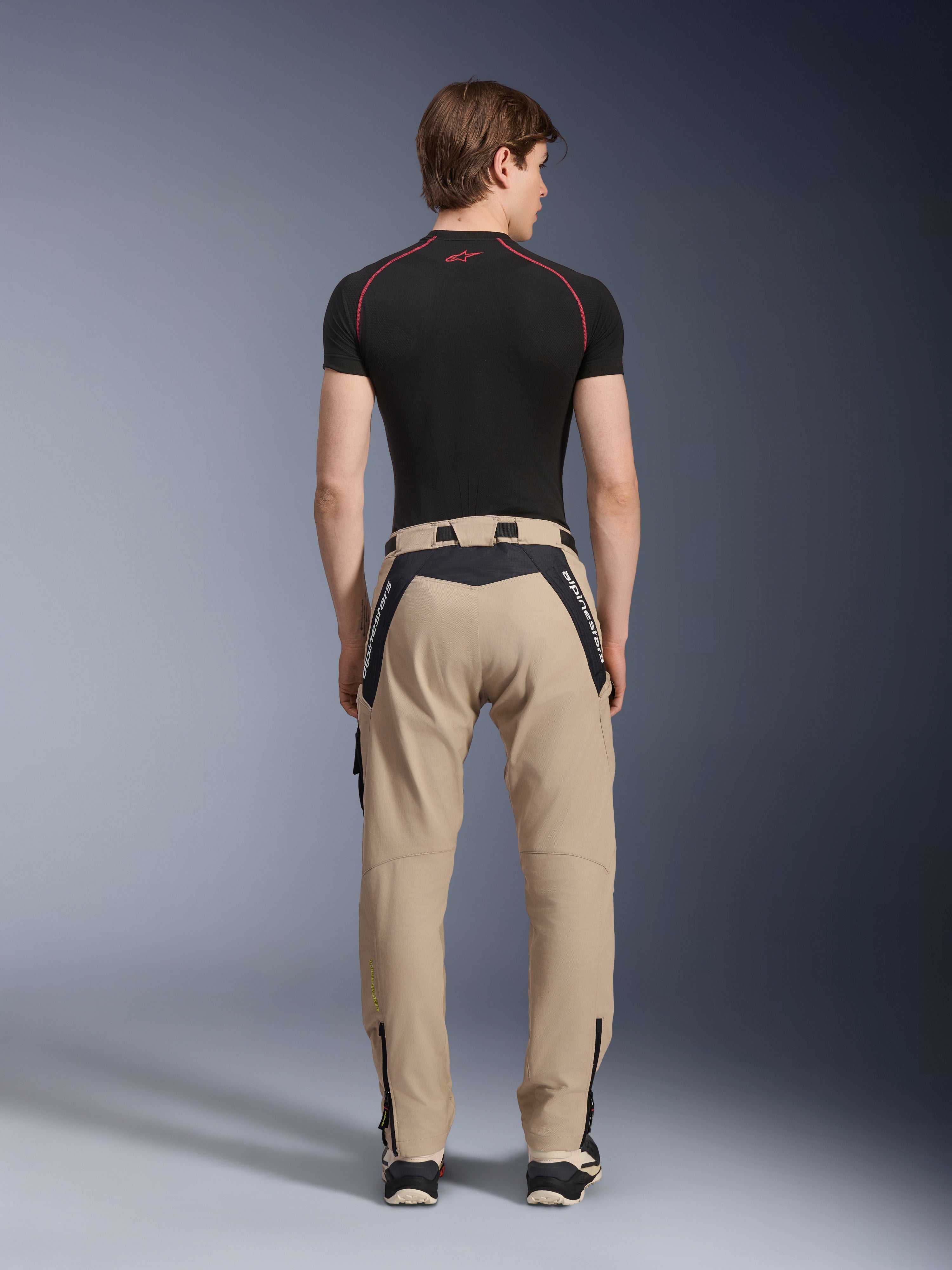 Alpinestars Acteon Pants in Sand, modern urban motorcycle pants viewed from the back, featuring black panels with white Alpinestars branding, tapered fit with zippers at the hem, worn with a black short-sleeved base layer and sneakers.