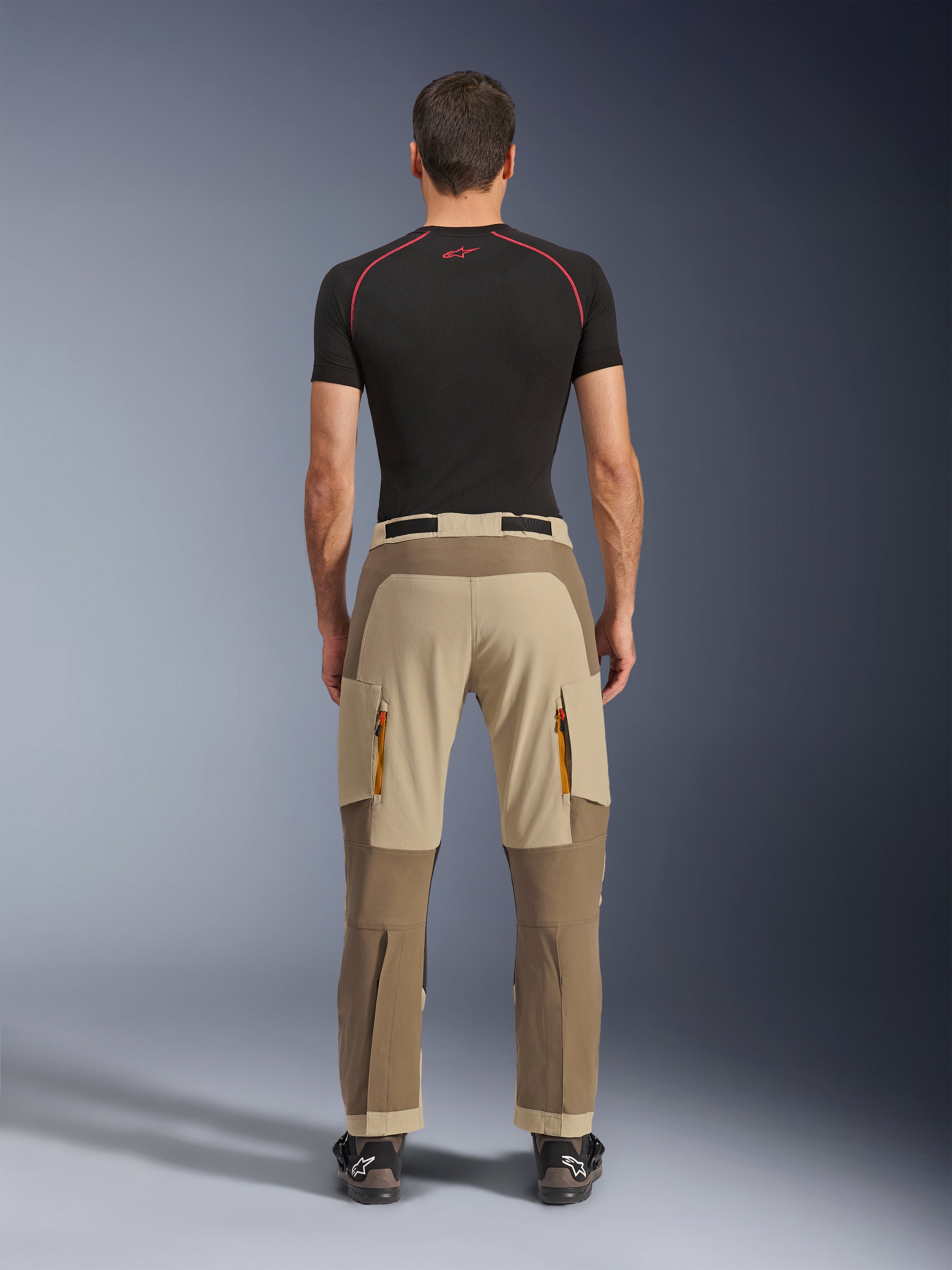 Alpinestars Maxdura Dual-byxor i Walnut Aluminum, sedda bakifrån, tvåtonad brun och beige design, äventyrsbyxor för motorcykelkörning med dragkedjeförsedda cargofickor och förstärkt säte, bärs med en svart kompressionströja och motorcykelstövlar.