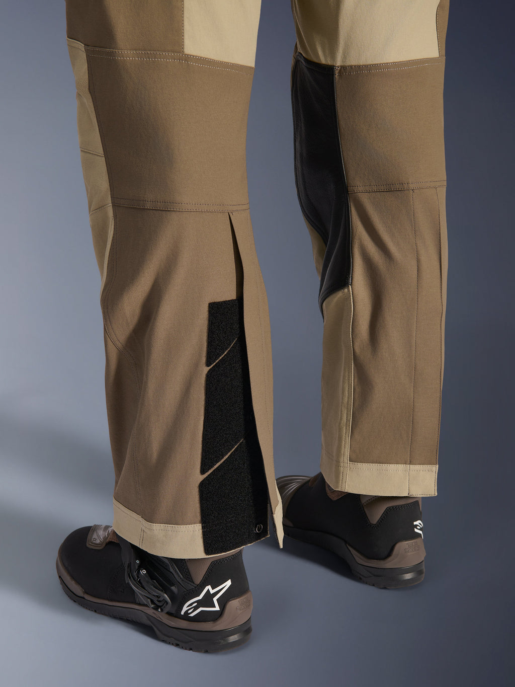 Alpinestars Maxdura Dual Pants i Walnut Aluminum, detaljerad vy av underben och nederkant med en over-the-boot-design, justerbara kardborrestängningar och förstärkta innerpaneler, matchade med svarta motorcykelstövlar.