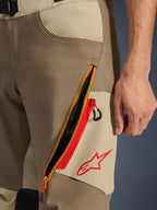 Alpinestars Maxdura Dual-byxor, Valnöt och Aluminium, närbild på lårdetalj med ett stort diagonalt ventilationssystem med dragkedja och röda accenter, röd Alpinestars-logotyp på en beige panel, äventyrsmotorcykelutrustning