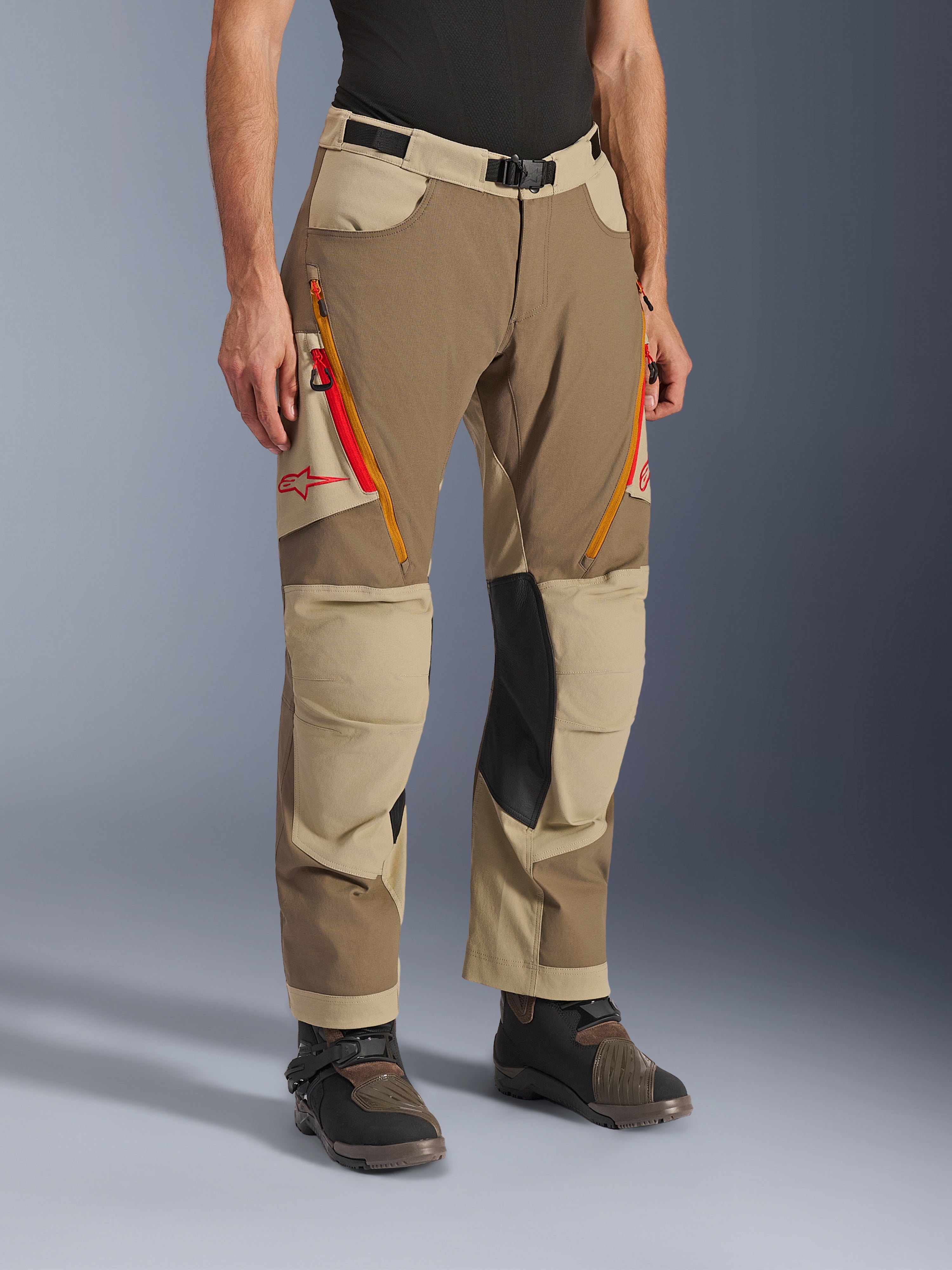 Alpinestars Maxdura Dual Pants, Walnut Aluminum bruna och khakifärgade äventyrsbyxor, med orangea och röda ventilationsöppningar med dragkedja på låren, förstärkta innerpaneler i svart läder vid knäna och röd Astars-logotyp, bärs med tekniska stövlar.