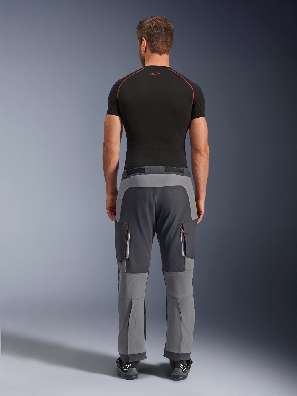 Alpinestars Maxdura Dual Pants, mörkgrå och djupgrå äventyrsmotorcykelbyxor, bakifrån på modell i svart t-shirt och stövlar, med grå paneler i flera nyanser och benfickor med dragkedja och röda detaljer.