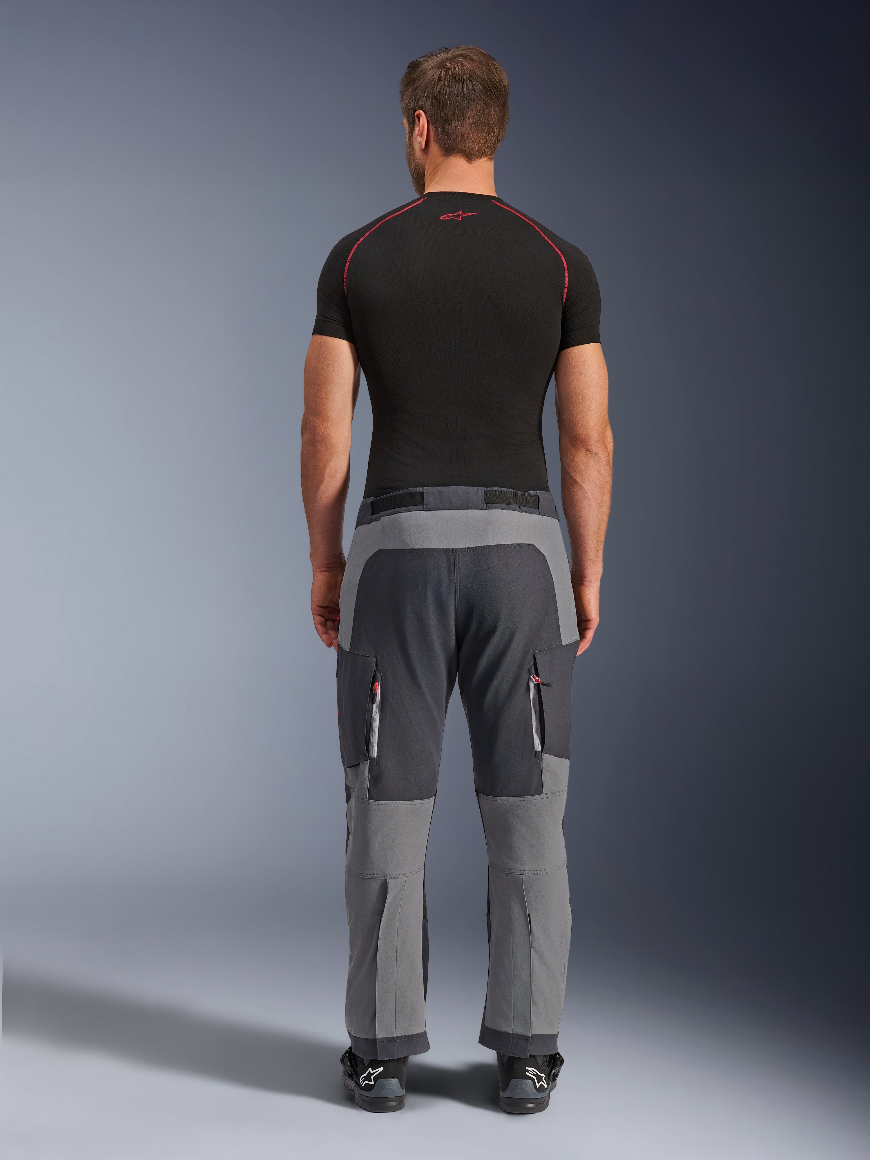 Alpinestars Maxdura Dual Pants, mörkgrå och djupgrå äventyrsmotorcykelbyxor, bakifrån på modell i svart t-shirt och stövlar, med grå paneler i flera nyanser och benfickor med dragkedja och röda detaljer.