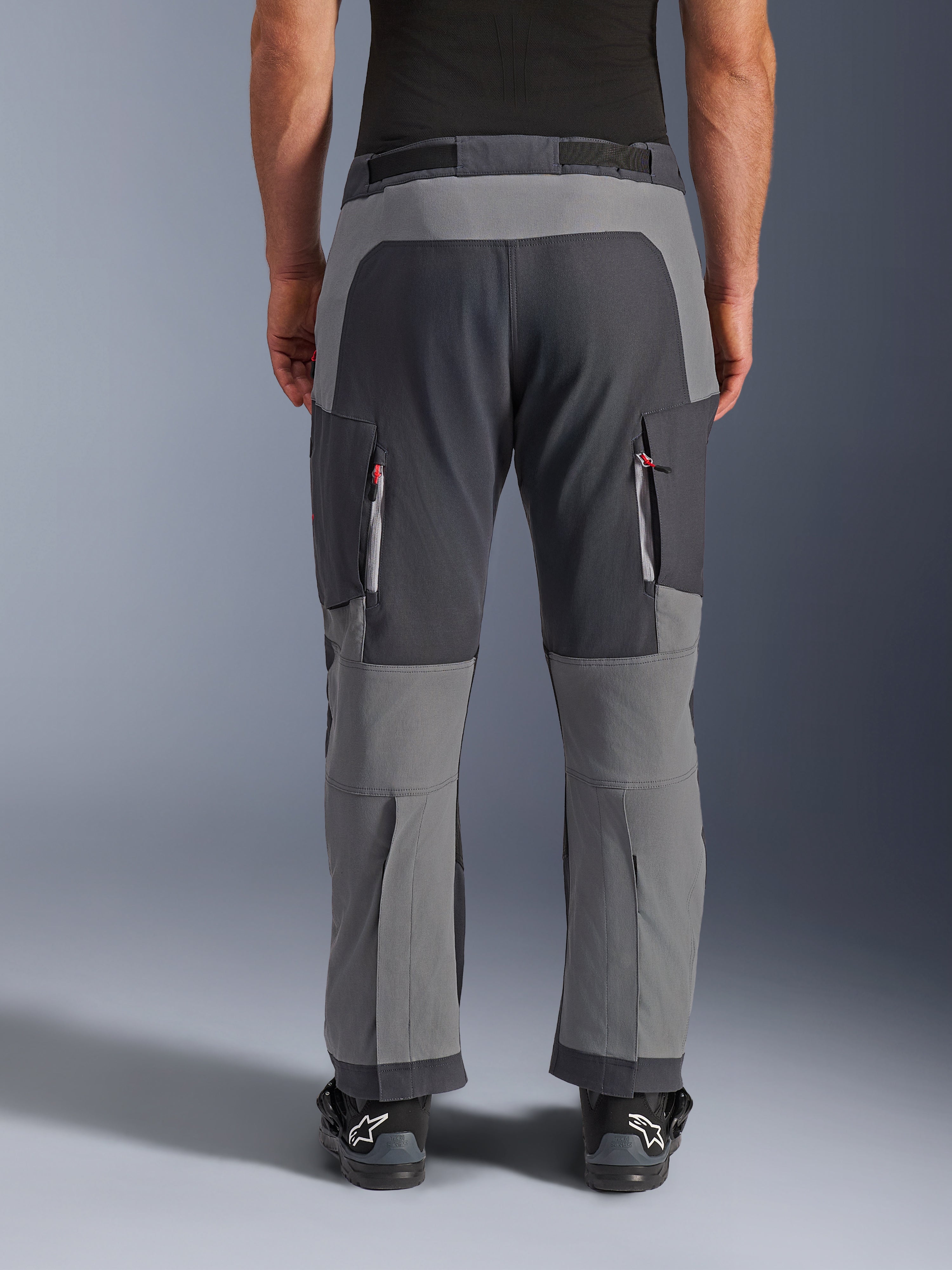 Alpinestars Maxdura Dual byxor, äventyrsmotorcykelbyxor, mörkgrå och djupgrå, vy bakifrån som framhäver förstärkta sätes- och knäpaneler, ventilationsöppningar med dragkedja på låren, bärs med svarta Alpinestars-stövlar, robust konstruktion i stretch-polyamid för dual-sport-körning