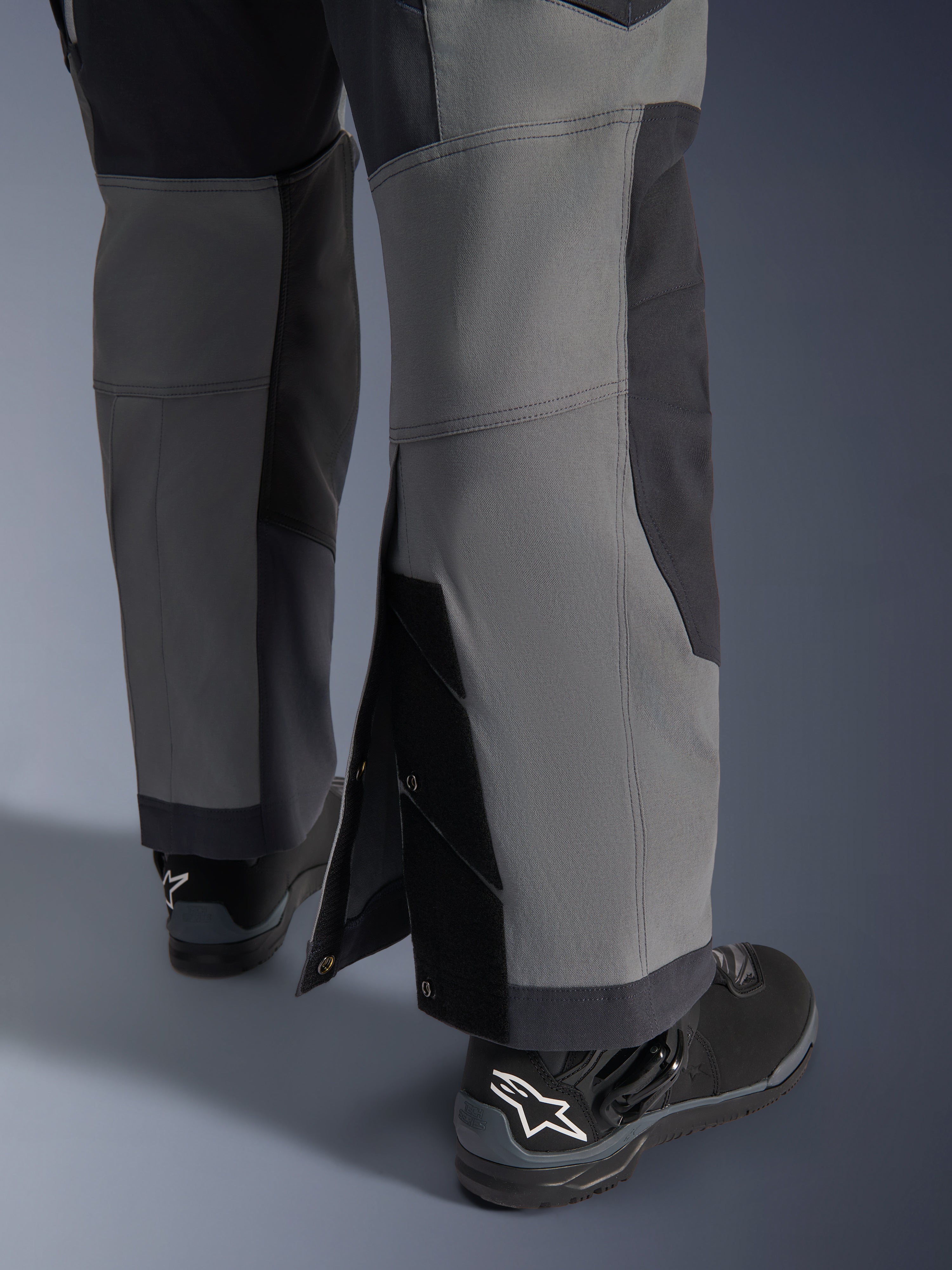 Alpinestars Maxdura Dual-byxor, äventyrsmotorcykelbyxor, mörkgrå och djupgrå, närbild av justerbara benslut som bärs över stövlarna med tryckknappar och förstärkta paneler, bärs med svarta Alpinestars-stövlar, robust teknisk design för dual-sport-körning