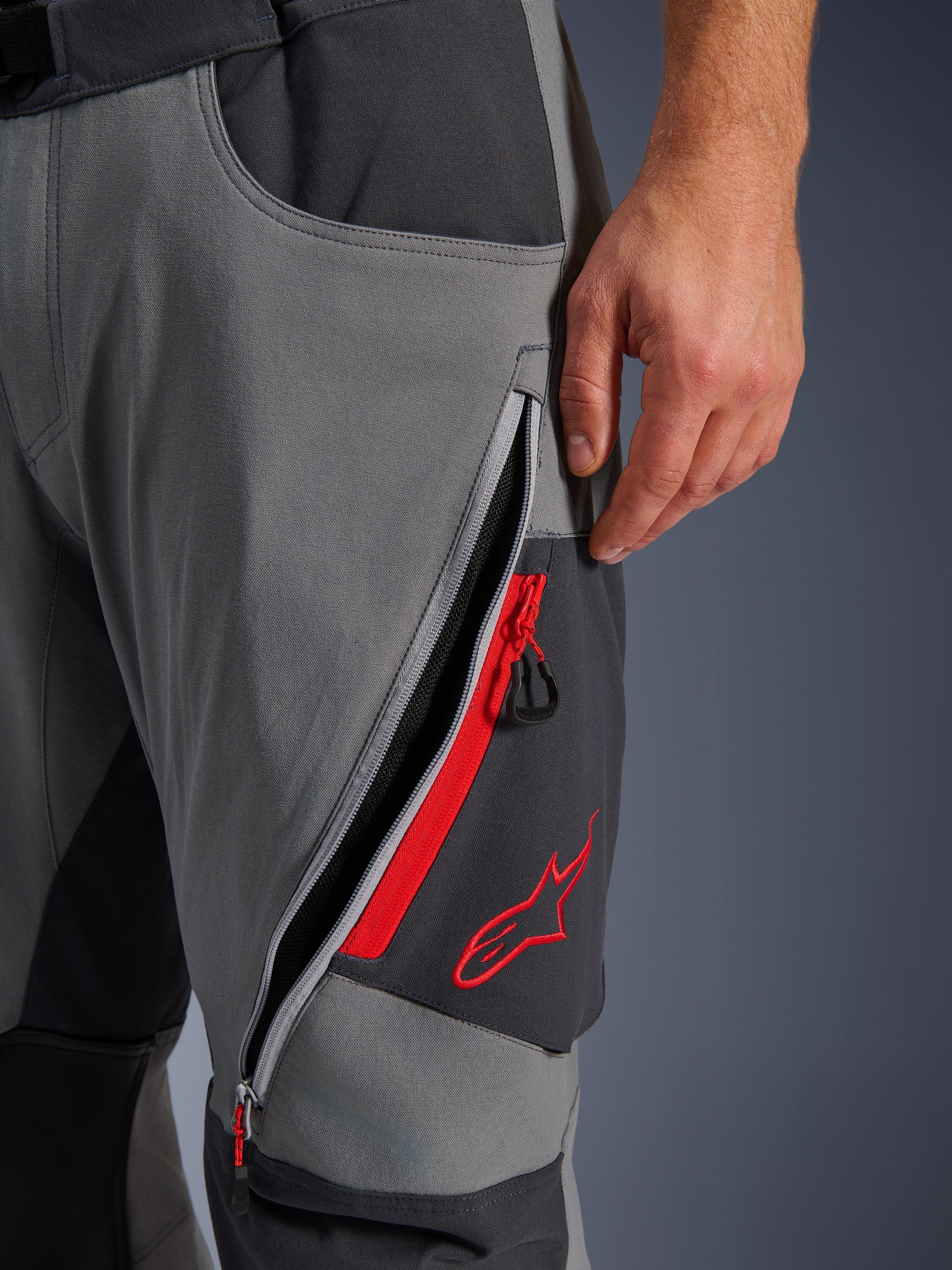 Alpinestars Maxdura Dual byxor, mörkgrå och djupgrå äventyrsbyxor för motorcykel, närbild på öppen lårventilation med dragkedja, röda detaljer och broderad röd stjärnlogotyp, teknisk stretchvävsdesign