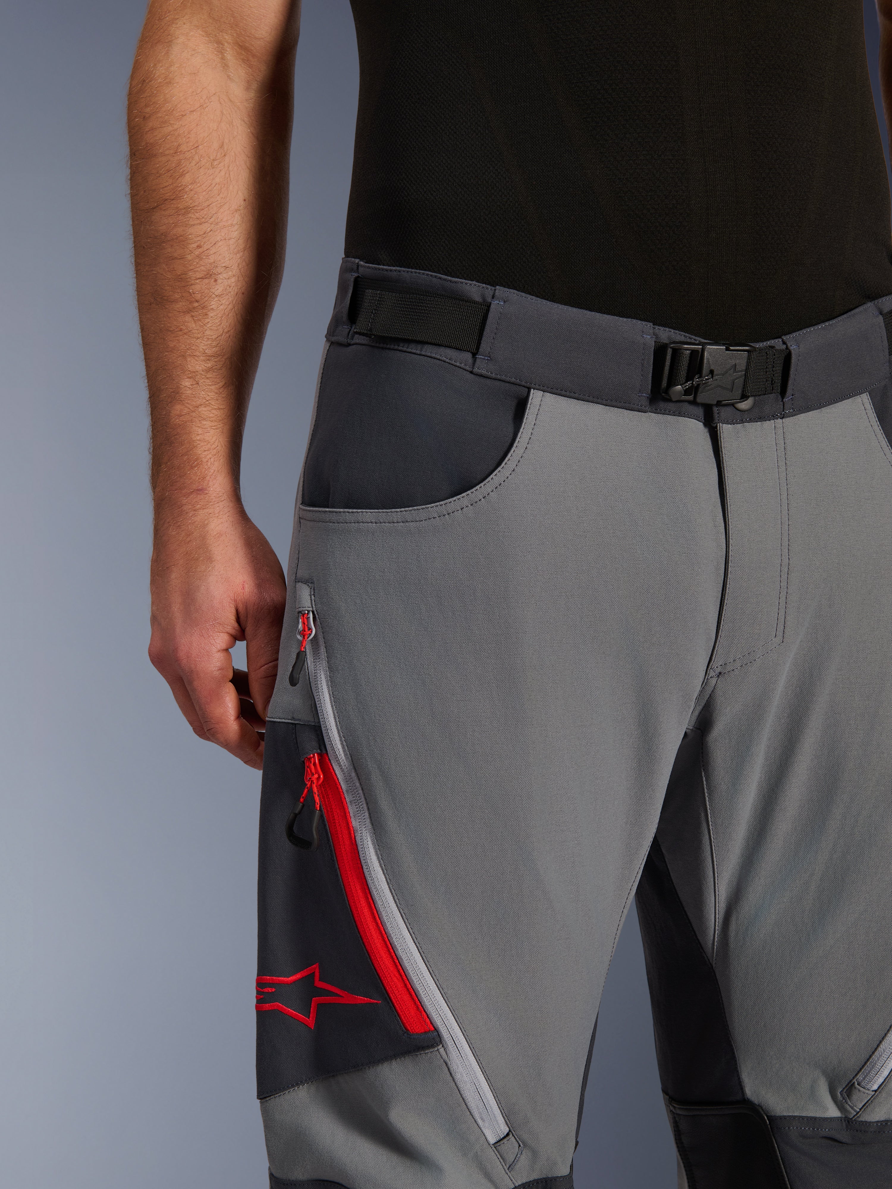 Alpinestars Maxdura Dual Pants, adventure-motorcykelbyxor, mörkgrå och djupgrå, närbild som visar justerbart midjebälte med magnetspänne, benficka med dragkedja med röda detaljer och Alpinestars-logotyp.