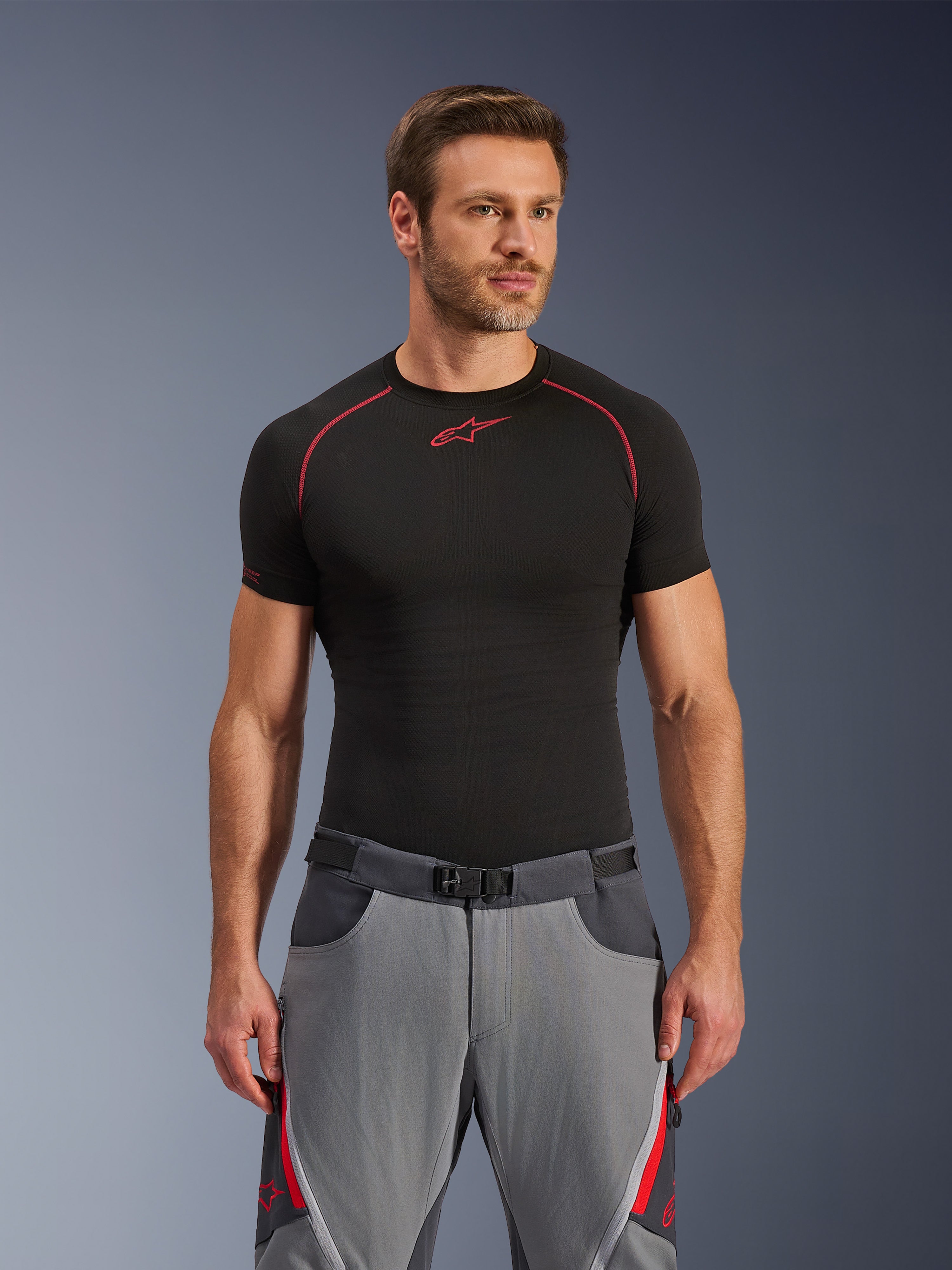 Alpinestars Maxdura Dual byxor, mörkgrå och djupgrå med röda detaljer, bärs med en svart underställströja med röda sömmar, atletisk passform med integrerat bälte med magnetspänne, slitstark MC-utrustning för äventyrsåkning