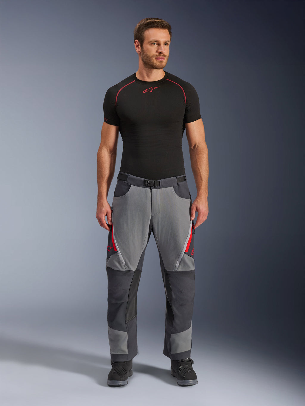 Alpinestars Maxdura Dual byxor, motorcykelbyxor, mörkgrå och djupgrå med röda detaljer, framifrån buren av en modell med ett svart Alpinestars-underställ och svarta stövlar, med förstärkta paneler och ventilationsöppningar med dragkedja på låren, körutrustning i äventyrsstil
