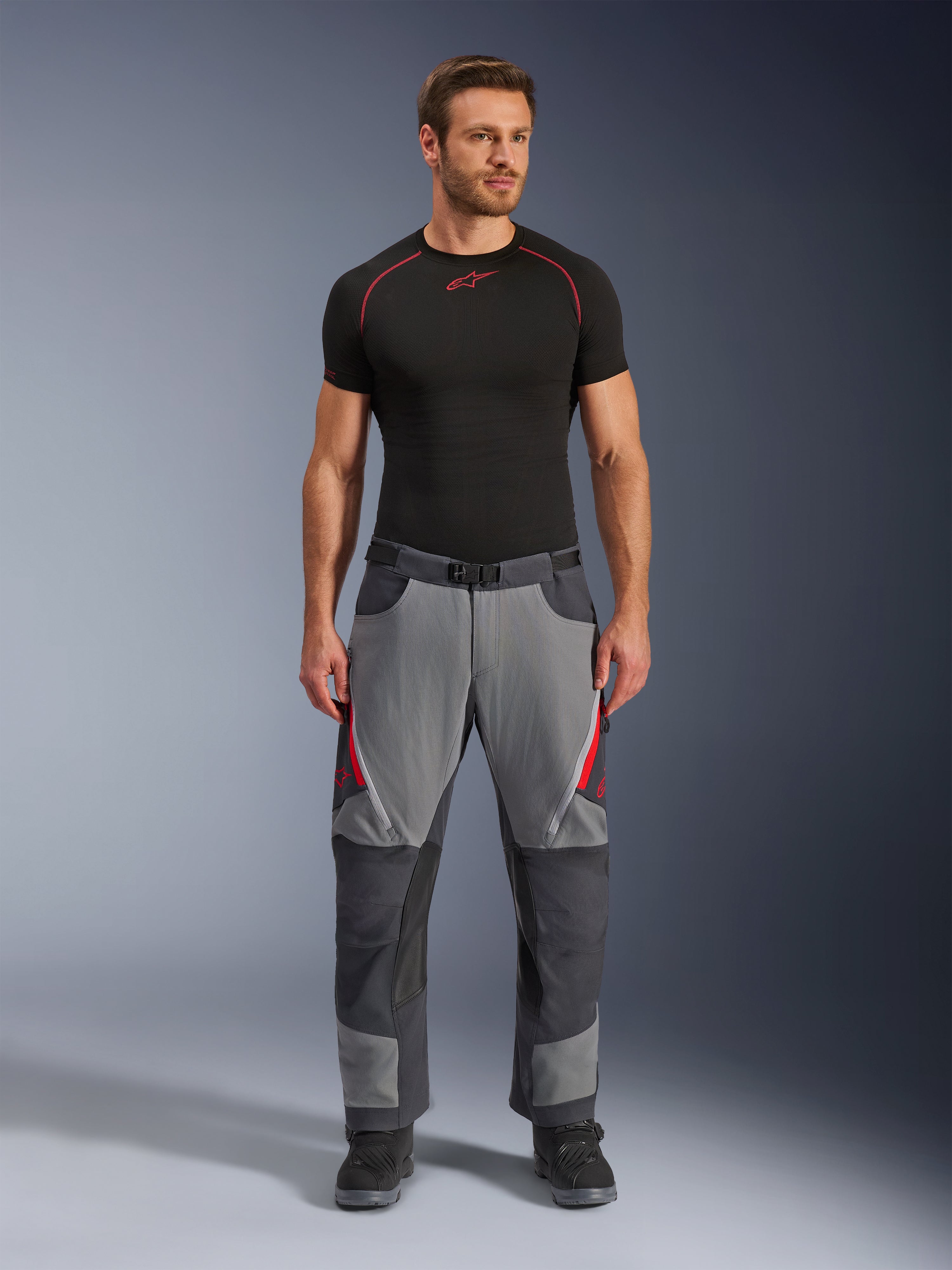 Alpinestars Maxdura Dual byxor, motorcykelbyxor, mörkgrå och djupgrå med röda detaljer, framifrån buren av en modell med ett svart Alpinestars-underställ och svarta stövlar, med förstärkta paneler och ventilationsöppningar med dragkedja på låren, körutrustning i äventyrsstil