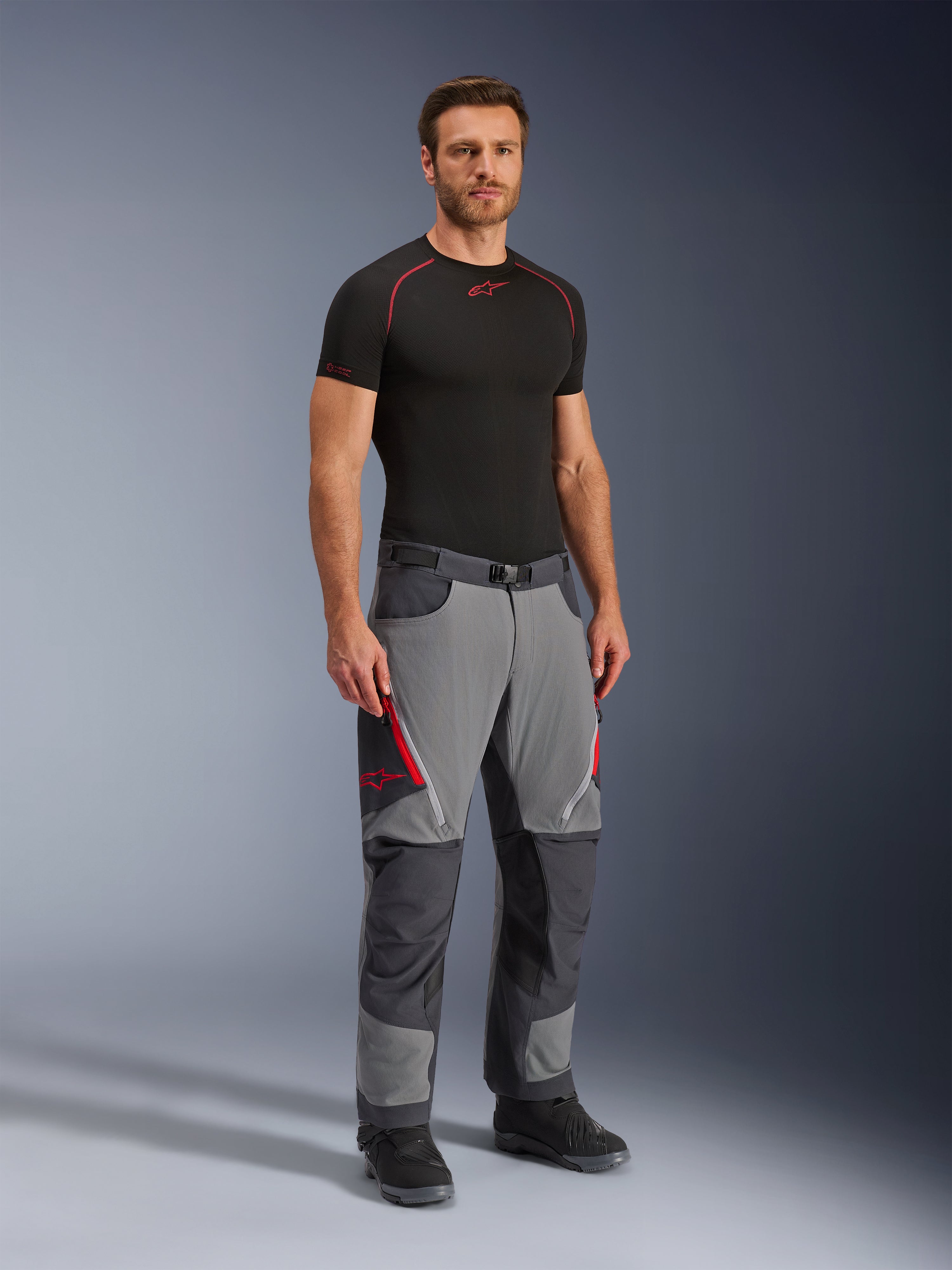 Alpinestars Maxdura Dual byxor, äventyrsmotorcykelbyxor, mörkgrå och djupgrå, bärs med ett svart underställ och stövlar, med röda dragkedjeförsedda ventiler, förstärkta paneler på knän och säte samt midja med bälte, atletisk passform för dual-sport-körning