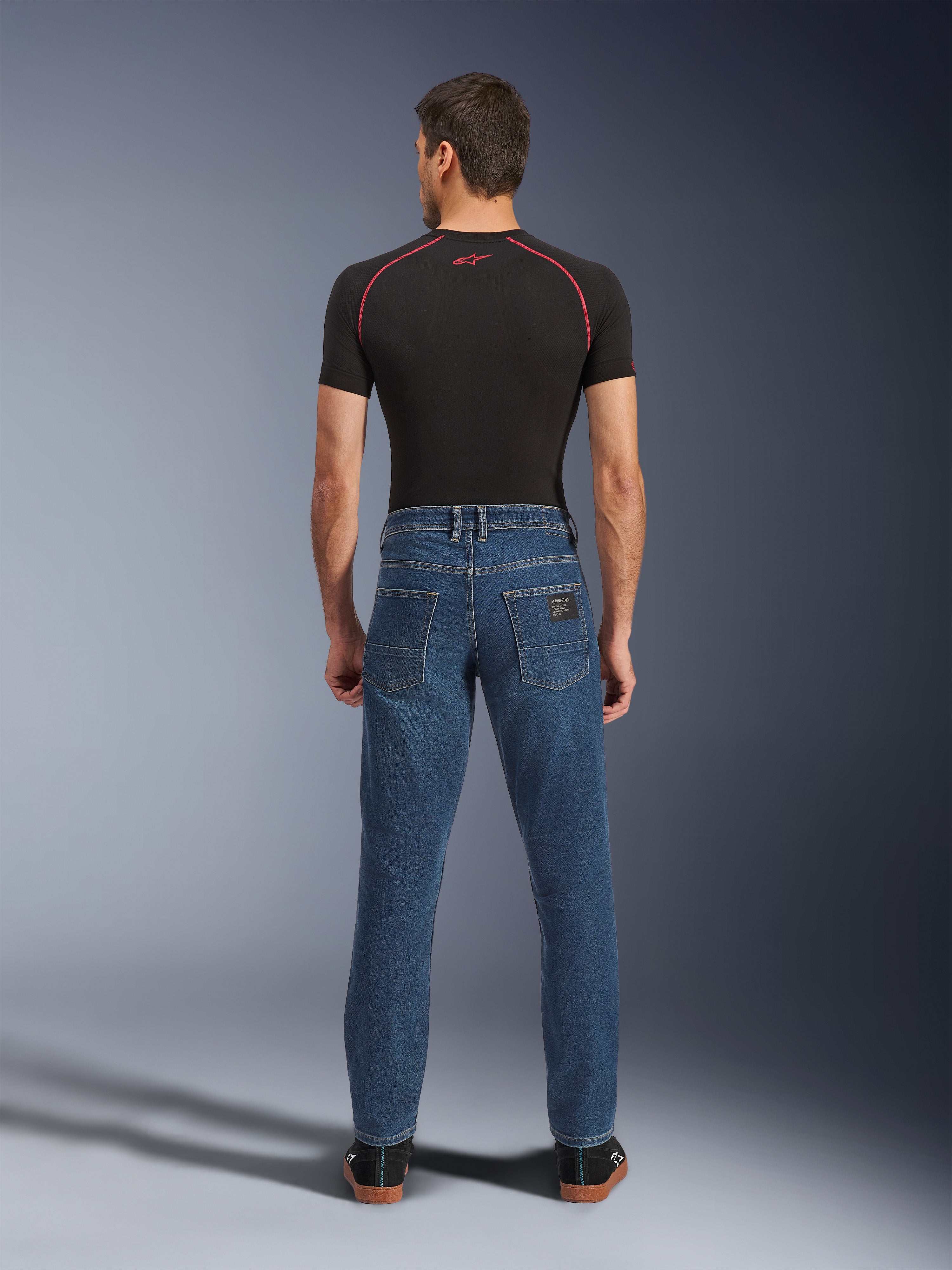 Alpinestars Flex-AST 5 Denim-byxor, Indigo Washed-blå, vy bakifrån som visar avsmalnande urban passform, bärs av modell med svart funktionströja och sneakers, skyddande MC-kläder i vardaglig stil