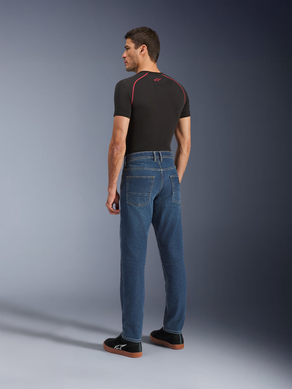 Alpinestars Flex-AST 5 Denim Pants, Indigo Washed, urbana motorcykelbyxor med avsmalnande passform, sedda bakifrån på en modell i svart t-shirt och sneakers, med en ren femficksdesign och stretchkonstruktion i ett lager