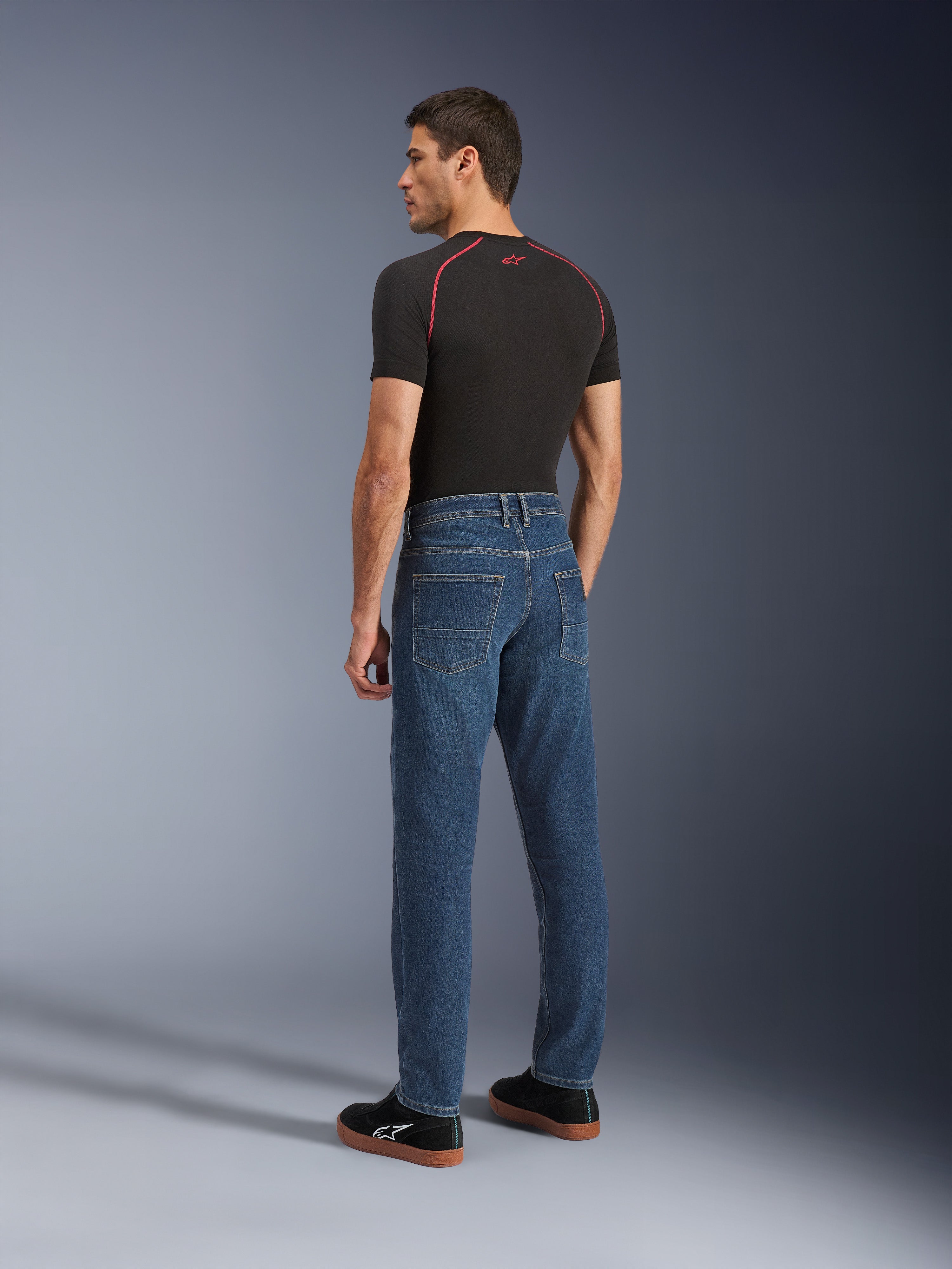 Alpinestars Flex-AST 5 Denim Pants, Indigo Washed, urbana motorcykelbyxor med avsmalnande passform, sedda bakifrån på en modell i svart t-shirt och sneakers, med en ren femficksdesign och stretchkonstruktion i ett lager