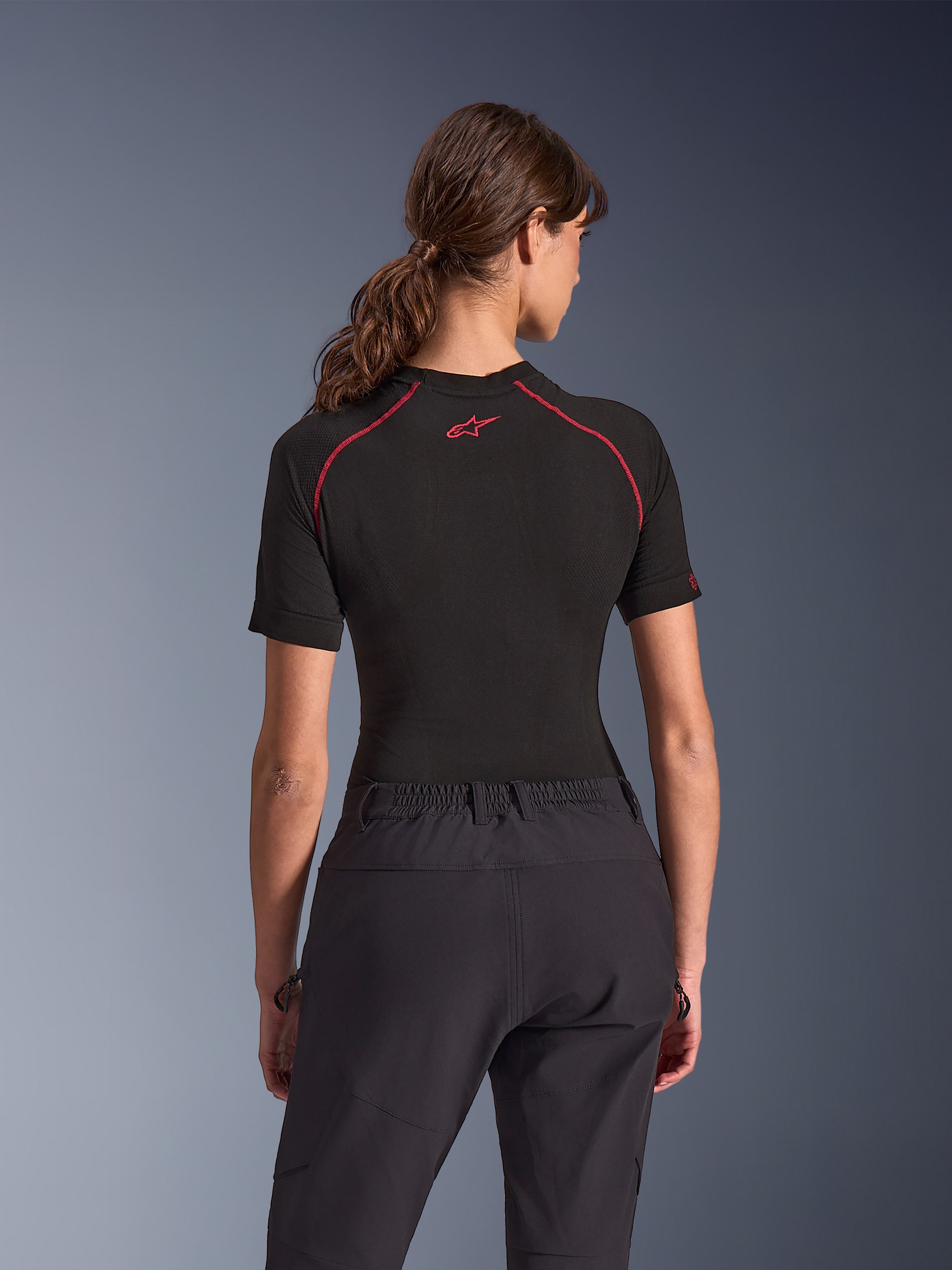 Kvinna i Alpinestars Stella Flex-AST Explorer Pants, svarta mc-byxor för äventyr, vy bakifrån, kombinerat med en svart kortärmad topp med röda sömmar, som framhäver den andningsbara textilkonstruktionen och det elastiska midjebandet.