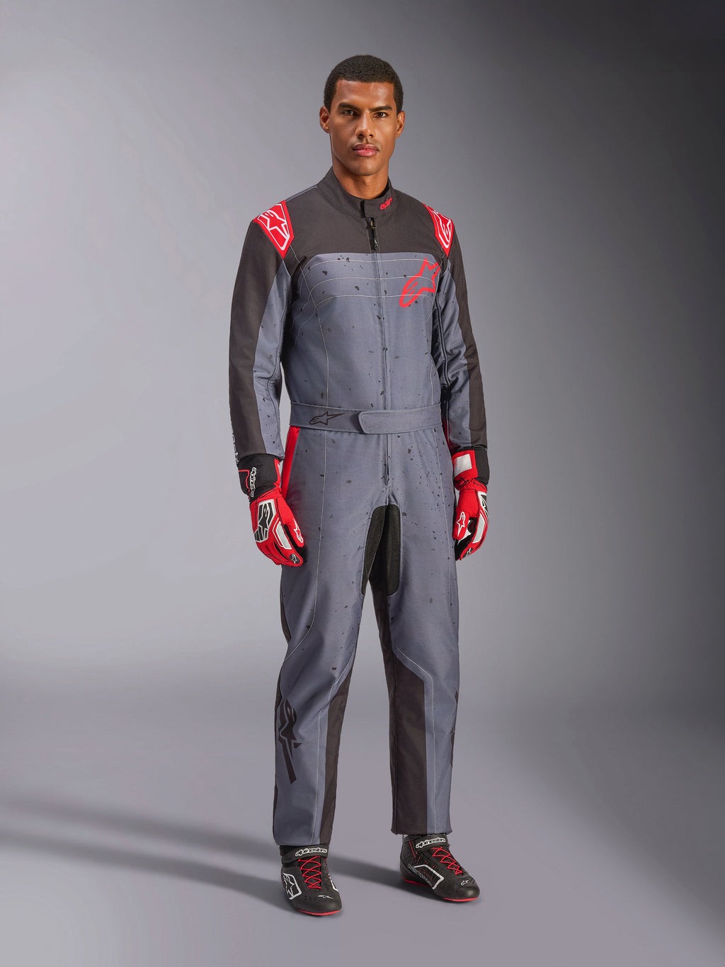 Alpinestars KMX-9 V3 Graphic 6 kartingdräkt, grå och antracit med röda detaljer, med ett unikt stänkmönster på bröstet och benen, matchat med röda racinghandskar och svarta kartingskor, helkroppsbild i studiomiljö.