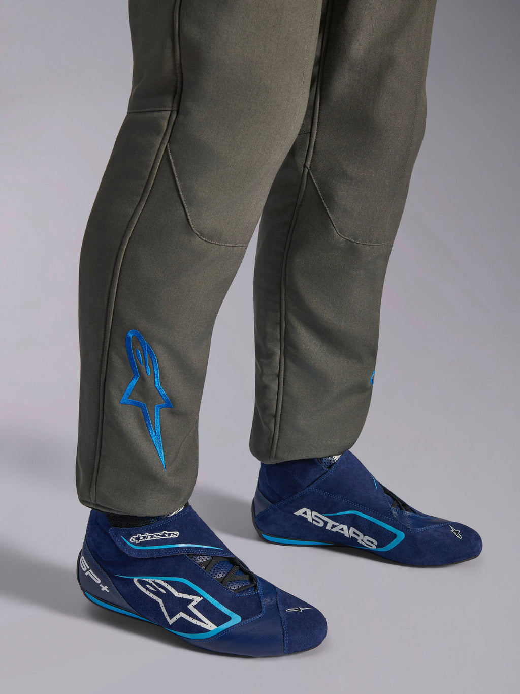 Alpinestars GP Race V3 Suit FIA, antracitblå racingoverall, närbild av underbenen med en blå broderad Astars-logotyp vid ankeln, matchad med marinblå och cyanfärgade racingskor, professionell skyddsutrustning för motorsport