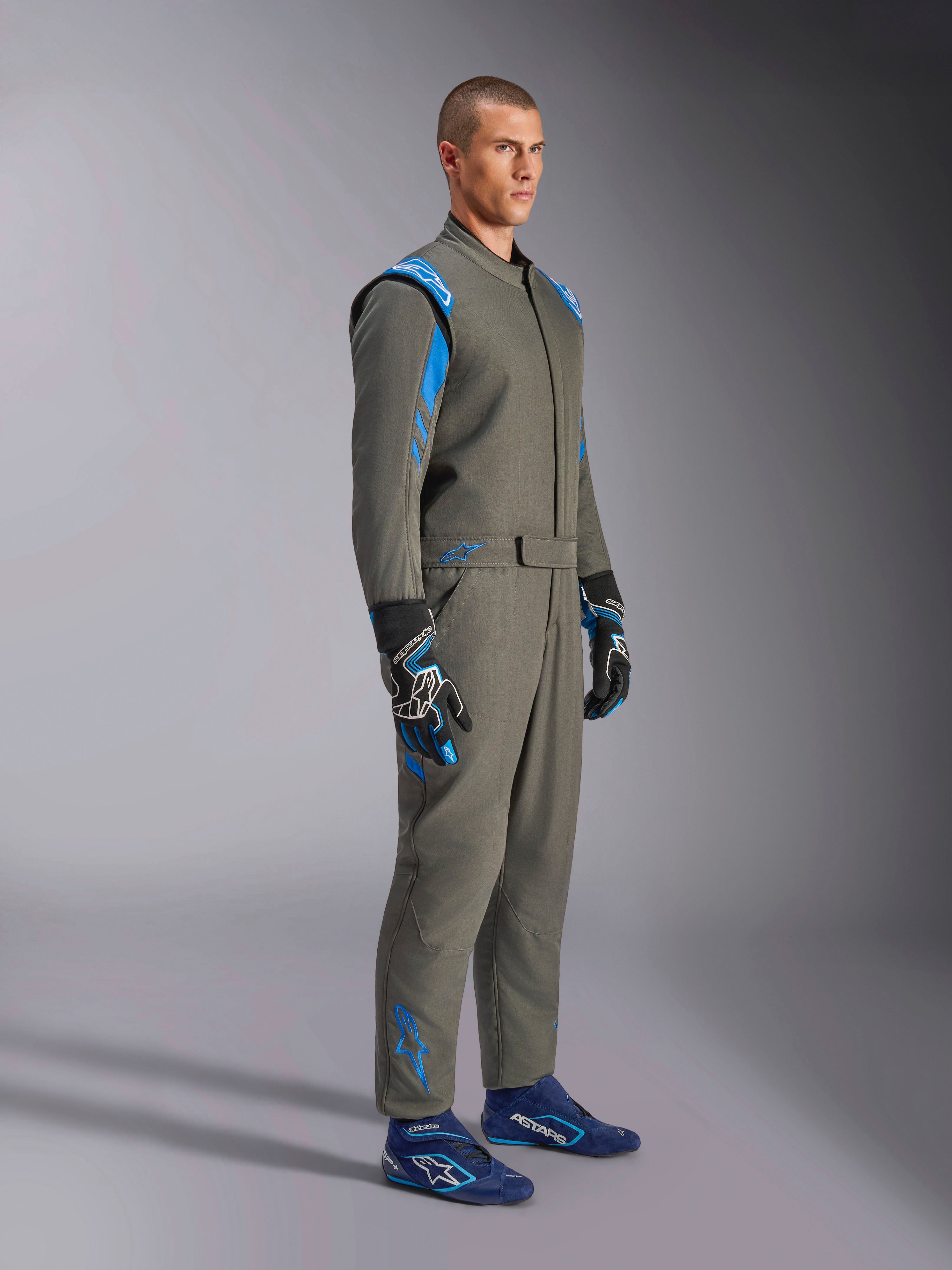 Alpinestars GP Race V3 Suit FIA i Anthracite Blue, racingoverall i ett stycke som bärs av en manlig modell, har klarblå accenter på axlar och anklar, kombinerad med matchande racinghandskar och blå racingskor, professionell motorsportutrustning.