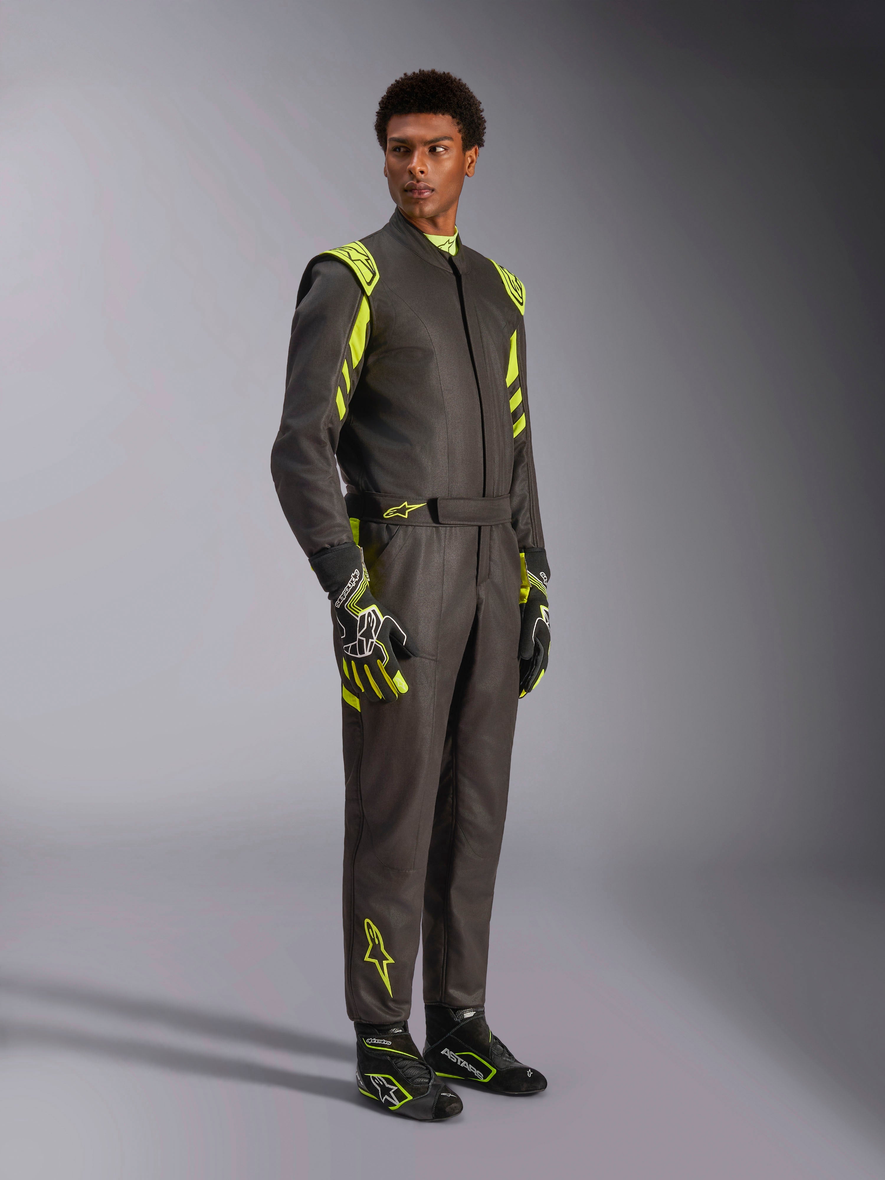 Alpinestars GP Race V3 Suit FIA, racingoverall för bilsport, svart och neongul, bärs av en modell med matchande racinghandskar och racingstövlar, iögonfallande neongula detaljer på axlar och ben, professionell motorsportutrustning