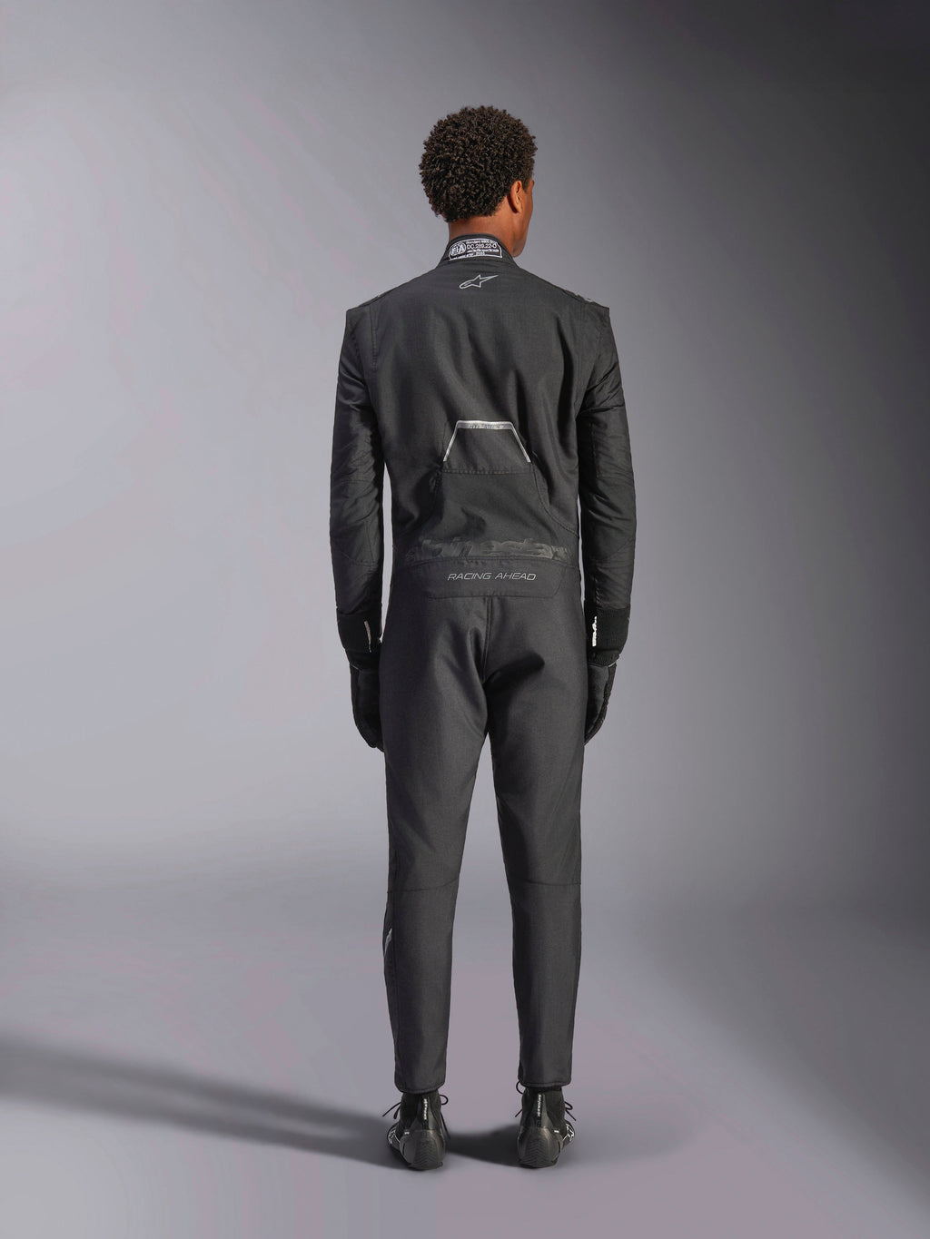 Alpinestars Hypertech V3 Suit, Black Dark Shadow, baksida av en professionell racingoverall i ett stycke för bilracing, med FIA-certifieringsetikett på kragen och "Alpinestars" och "RACING AHEAD"-märkning på ländryggen, bärs med matchande svarta racinghandskar och stövlar.
