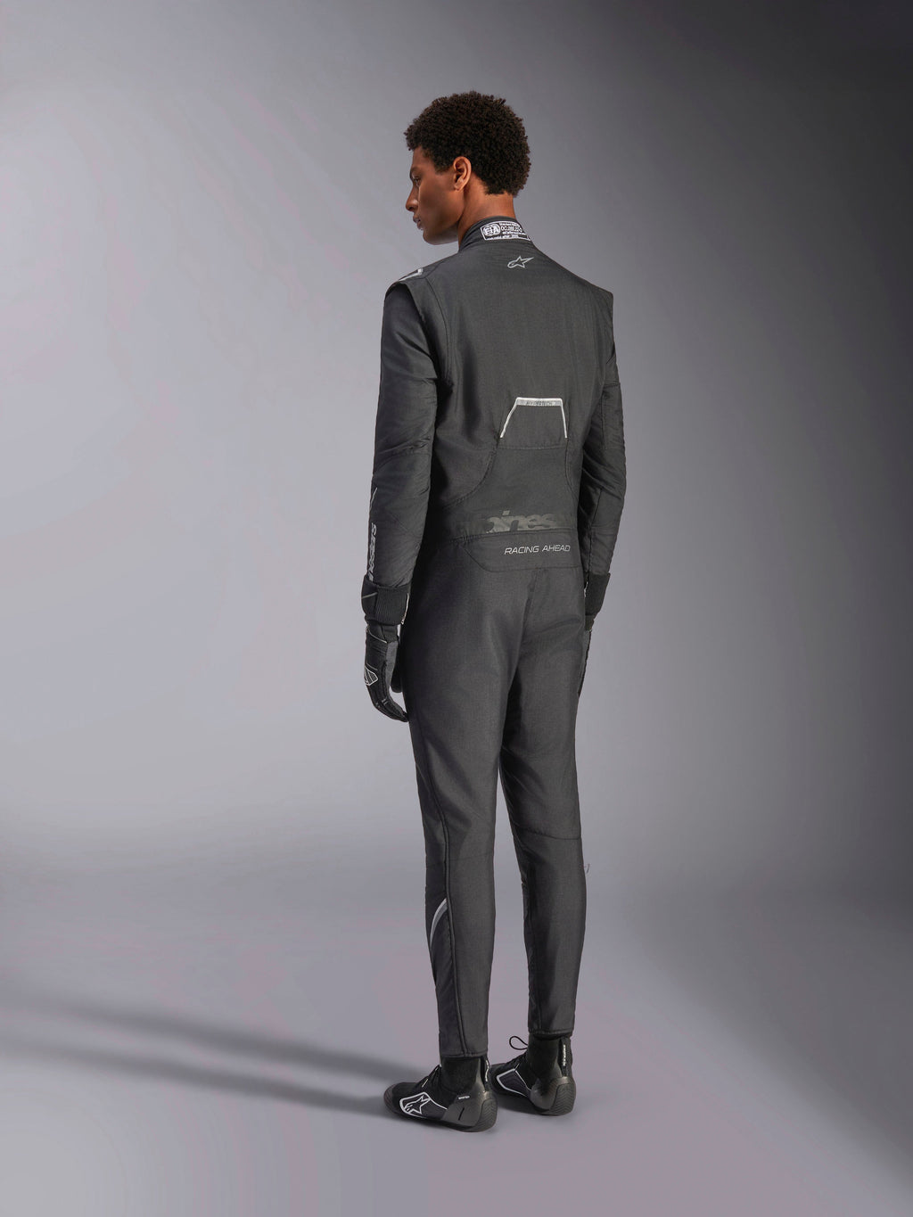 Alpinestars Hypertech V3 Suit, Black Dark Shadow, ryggvy av manlig modell klädd i professionell racingoverall med matchande svarta handskar och skor, lätt Nomex-konstruktion och FIA-certifiering på kragen, athletic-slim passform för motorsport.