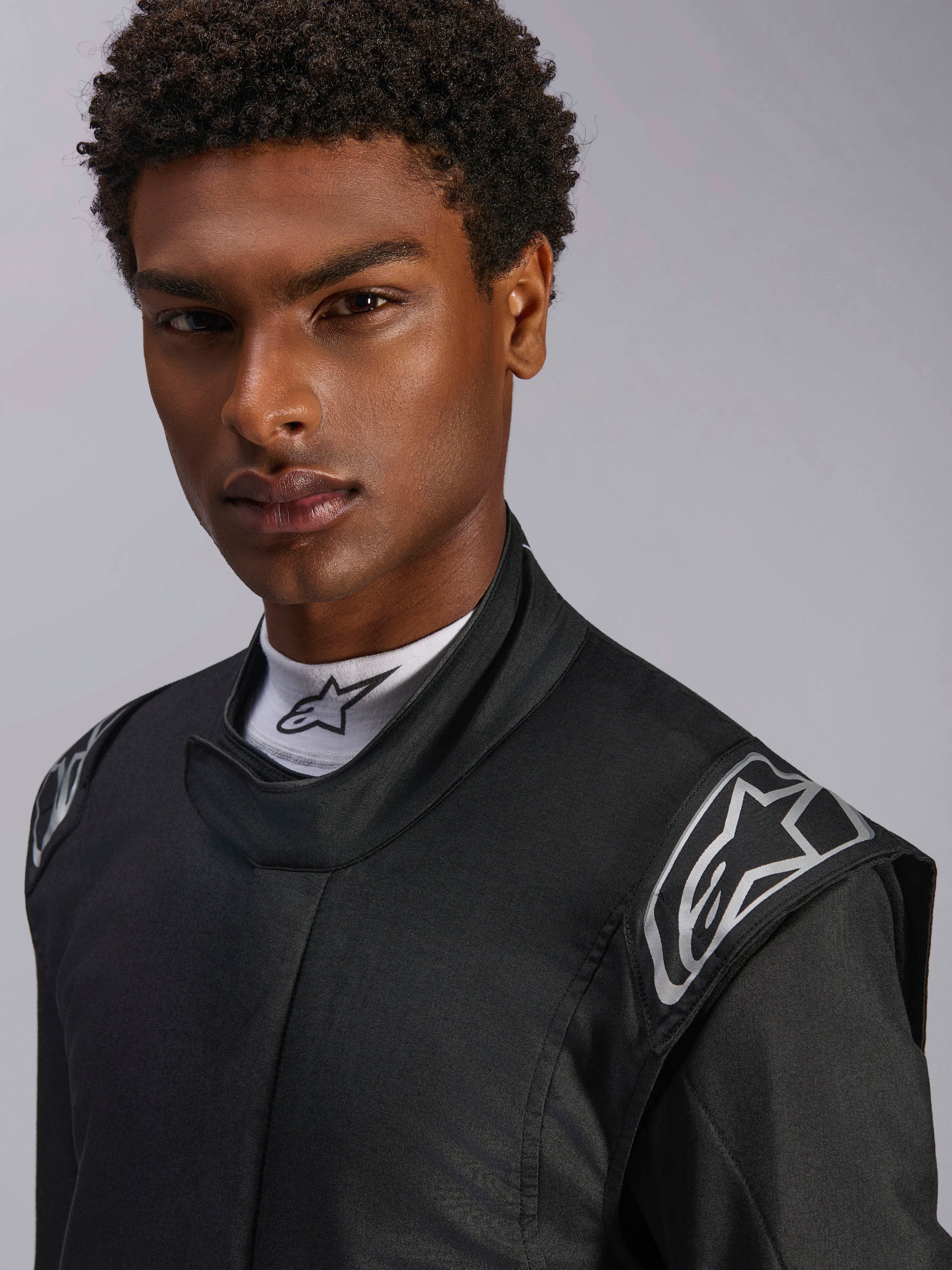 Alpinestars Hypertech V3 Suit, Black Dark Shadow racingoverall, närbild på manlig modell, silverfärgade axellogotyper, hög krage med vit Alpinestars mock-neck-tröja, professionell skyddsutrustning för motorsport