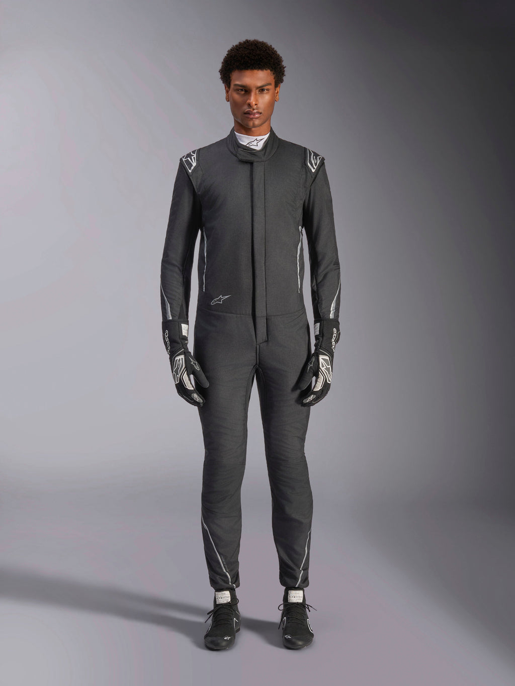 Alpinestars Hypertech V3 Suit, professionell racingoverall för bilsport, Black och Dark Shadow, bärs av en modell med matchande racinghandskar och skor, slimmad atletisk passform med silverdetaljer och logotyper, högpresterande motorsportutrustning