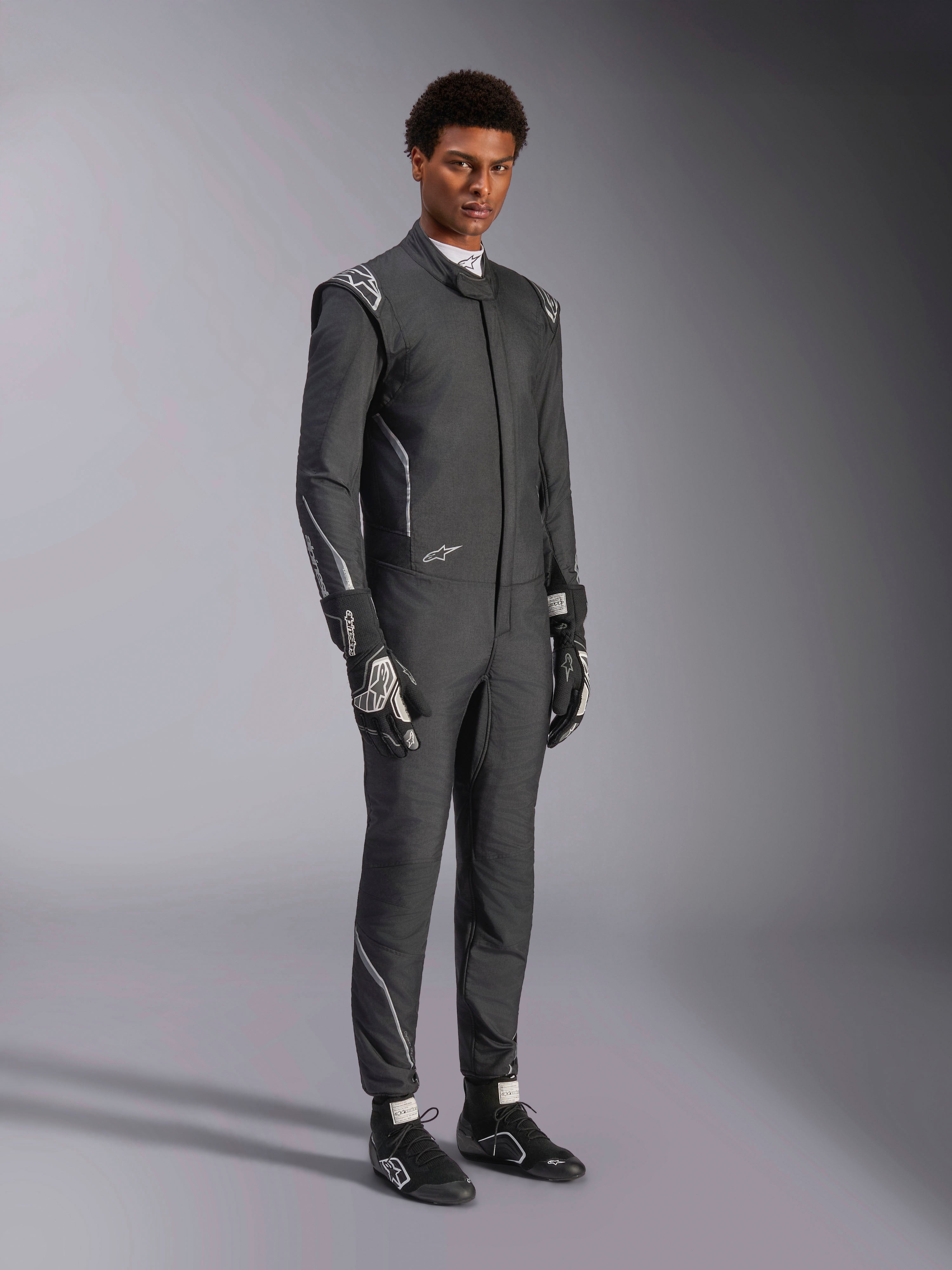 Alpinestars Hypertech V3-dräkt, racingoverall, svart och Dark Shadow, bärs av en manlig modell med matchande svarta handskar och racingskor, atletisk slim fit, har Alpinestars-logotyper och ljusgrå accenter, professionell skyddsutrustning för motorsport