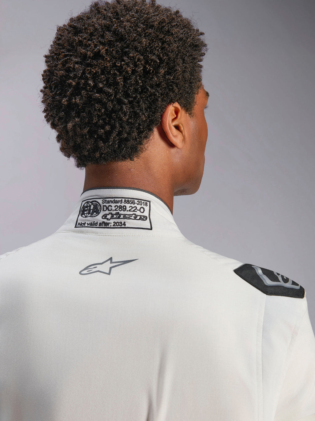 Alpinestars Hypertech V3-dräkt, vit, detaljvy av nackkrage och axel, med FIA 8856-2018-certifieringsetikett, tryckt grå Alpinestars-logotyp och svart evakueringshandtag, professionell racingutrustning