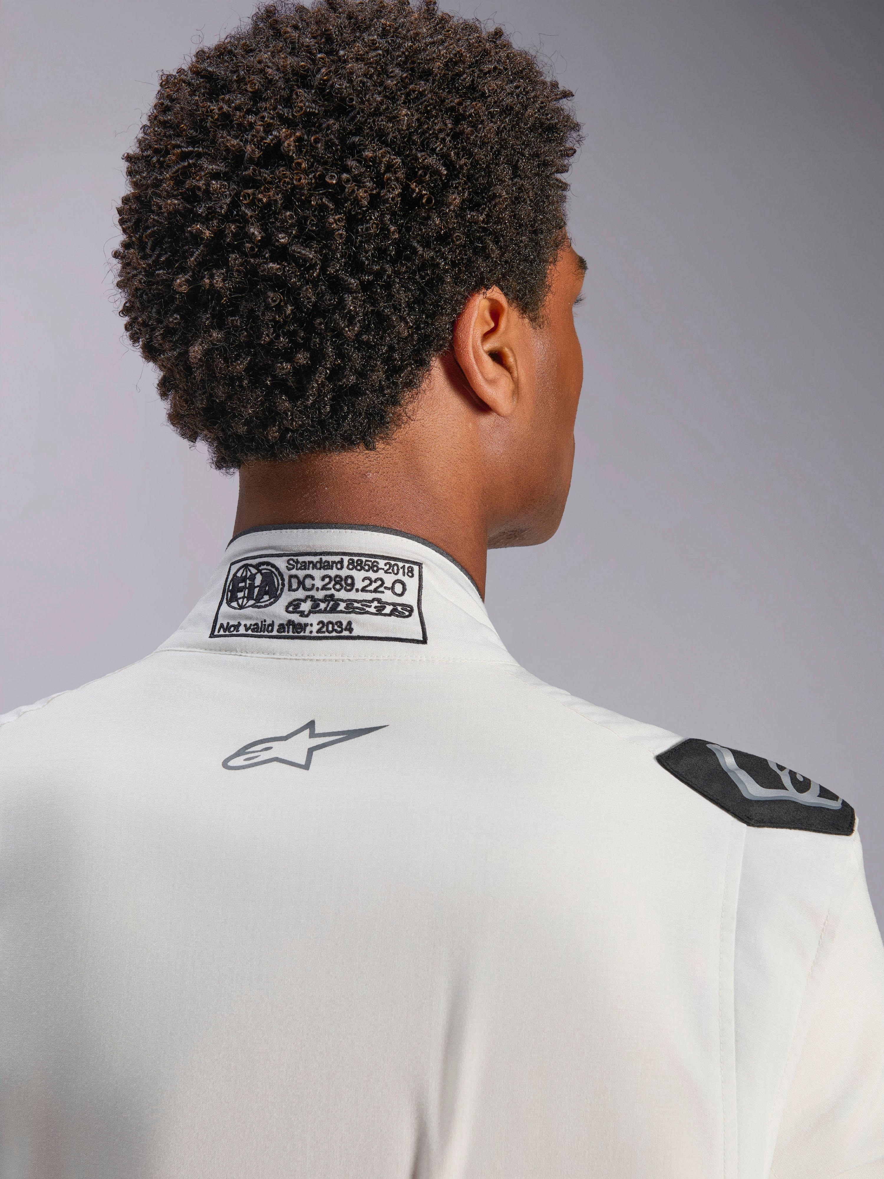 Alpinestars Hypertech V3-dräkt, vit, detaljvy av nackkrage och axel, med FIA 8856-2018-certifieringsetikett, tryckt grå Alpinestars-logotyp och svart evakueringshandtag, professionell racingutrustning