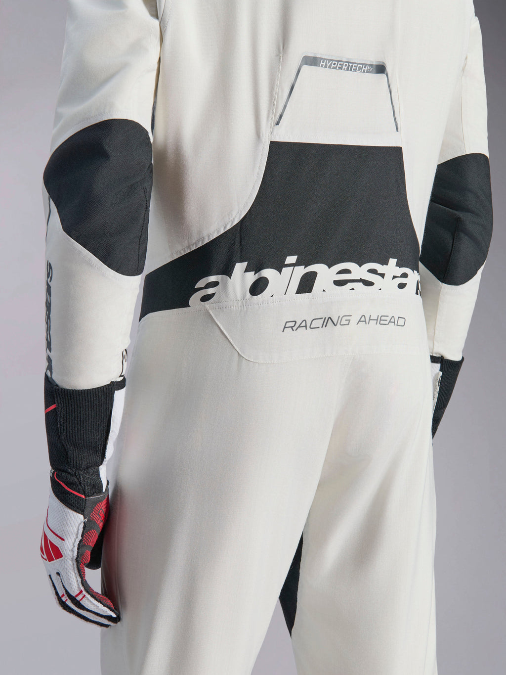 Alpinestars Hypertech V3-dräkt, vit, svart och silverfärgad racingoverall för bil, sedd bakifrån med stor svart 'alpinestars'-logotyp och 'RACING AHEAD'-text, svarta stretchpaneler på armbågar och ländrygg, professionell skyddsutrustning för motorsport.