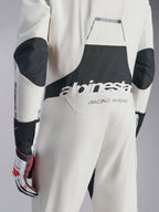 Alpinestars Hypertech V3-dräkt, vit, svart och silverfärgad racingoverall för bil, sedd bakifrån med stor svart 'alpinestars'-logotyp och 'RACING AHEAD'-text, svarta stretchpaneler på armbågar och ländrygg, professionell skyddsutrustning för motorsport.