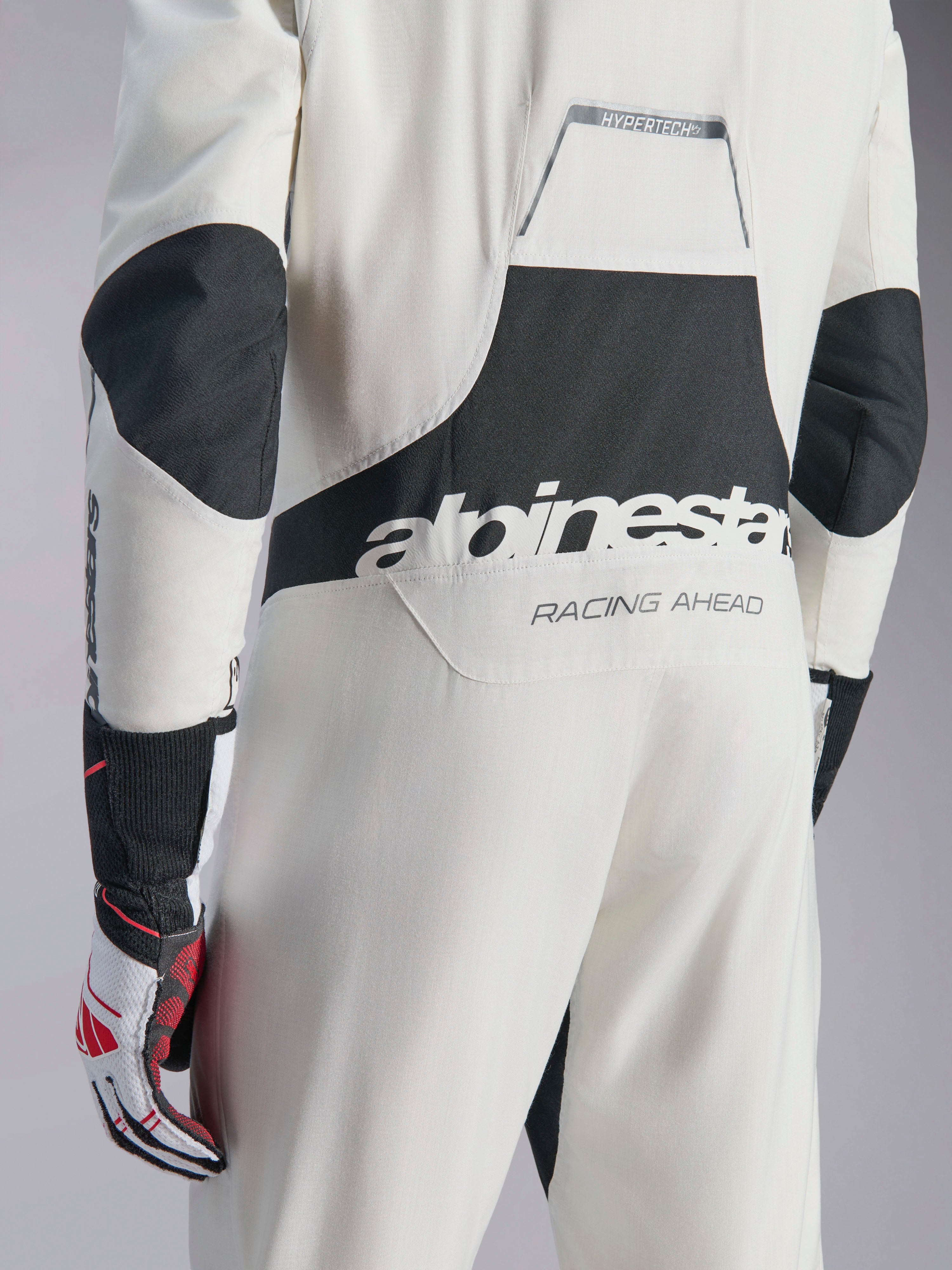 Alpinestars Hypertech V3-dräkt, vit, svart och silverfärgad racingoverall för bil, sedd bakifrån med stor svart 'alpinestars'-logotyp och 'RACING AHEAD'-text, svarta stretchpaneler på armbågar och ländrygg, professionell skyddsutrustning för motorsport.