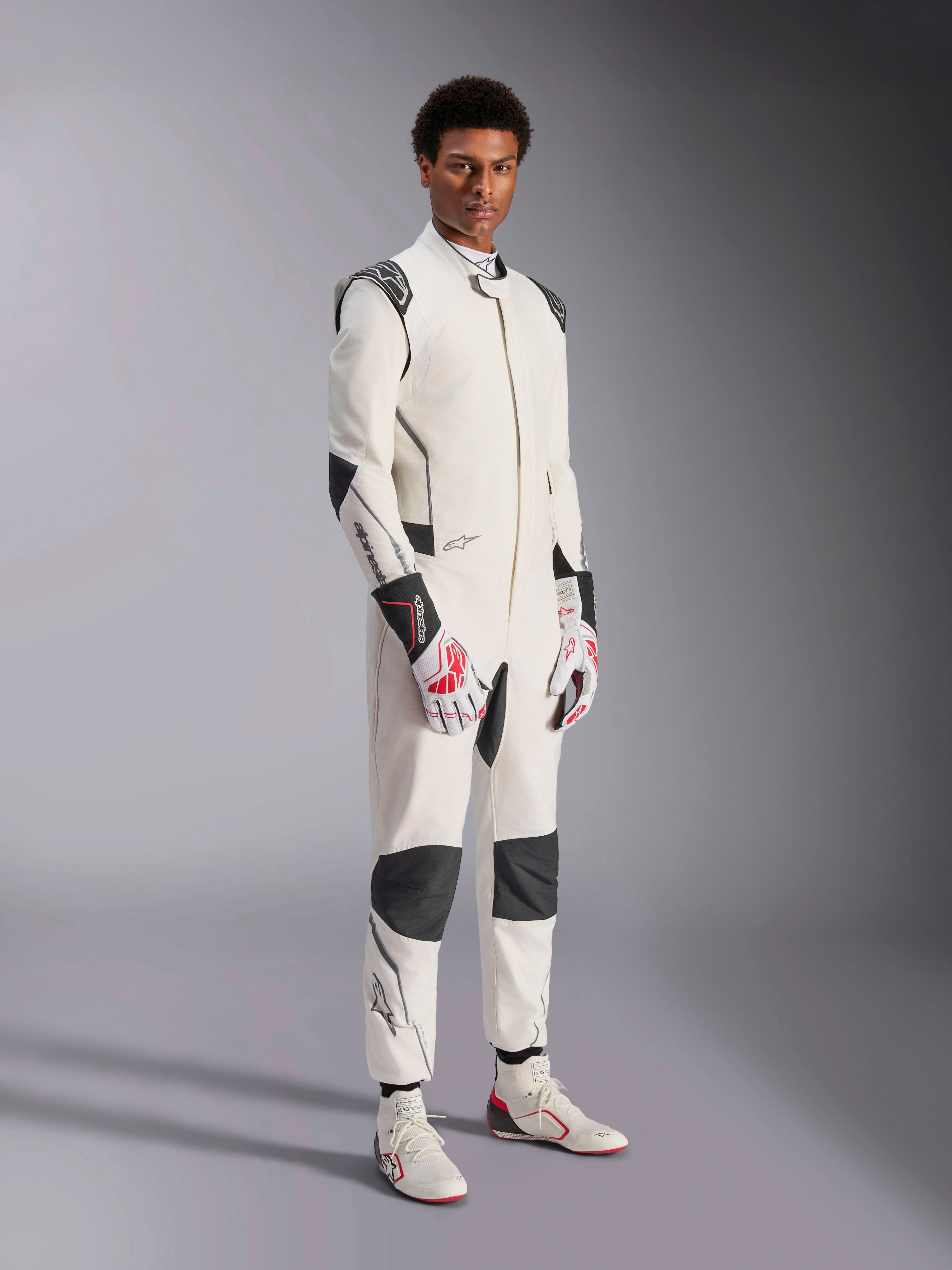 Manlig modell i Alpinestars Hypertech V3 Suit, vit med svarta och silvriga detaljer, professionell bilracingutrustning med svarta paneler på axlar och knän, matchad med vita och röda racinghandskar och vita racingstövlar