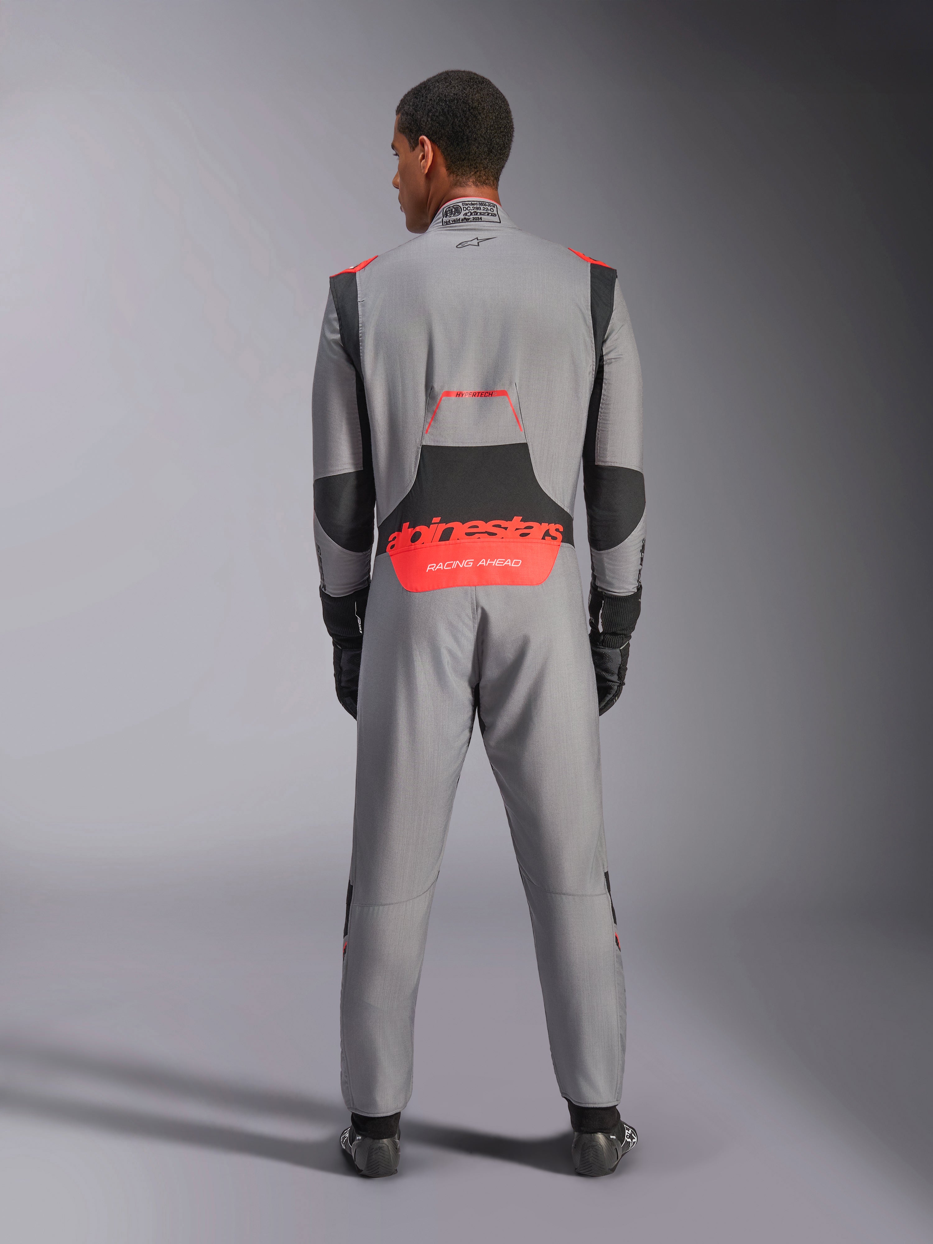 Alpinestars Hypertech V3-overall, professionell racingoverall för bil, mellangrå med svarta och röd fluo-detaljer, bakifrån på en modell med röd Alpinestars-logotyp på ländryggen, lätt Nomex-konstruktion för banprestanda.