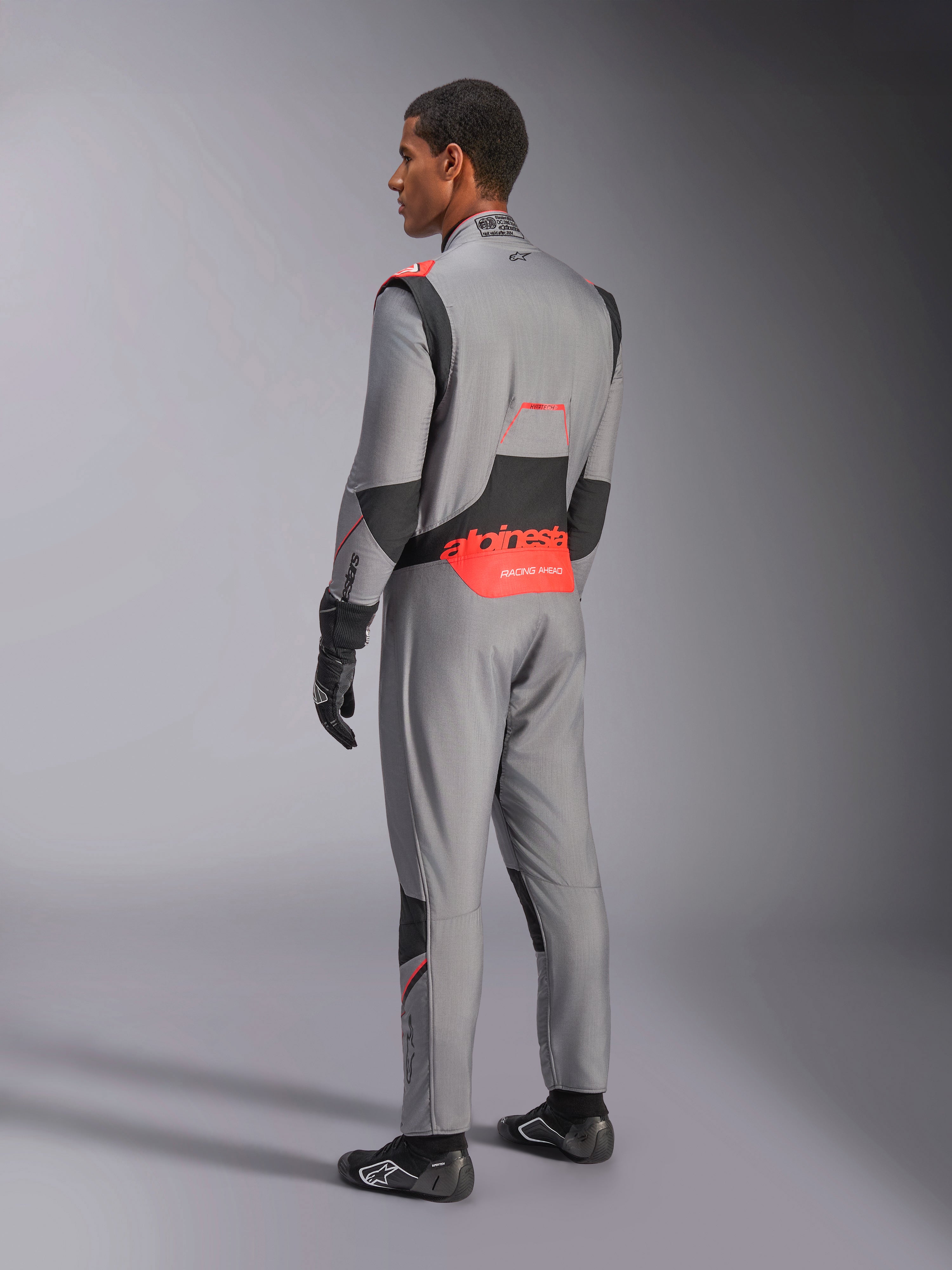 Alpinestars Hypertech V3-dräkt i Mid Gray Black Red Fluo, trekvartsvy bakifrån, med framträdande röd Alpinestars-logotyp på ländryggen, svarta stretchpaneler och en smidig atletisk passform för professionell bilsport.