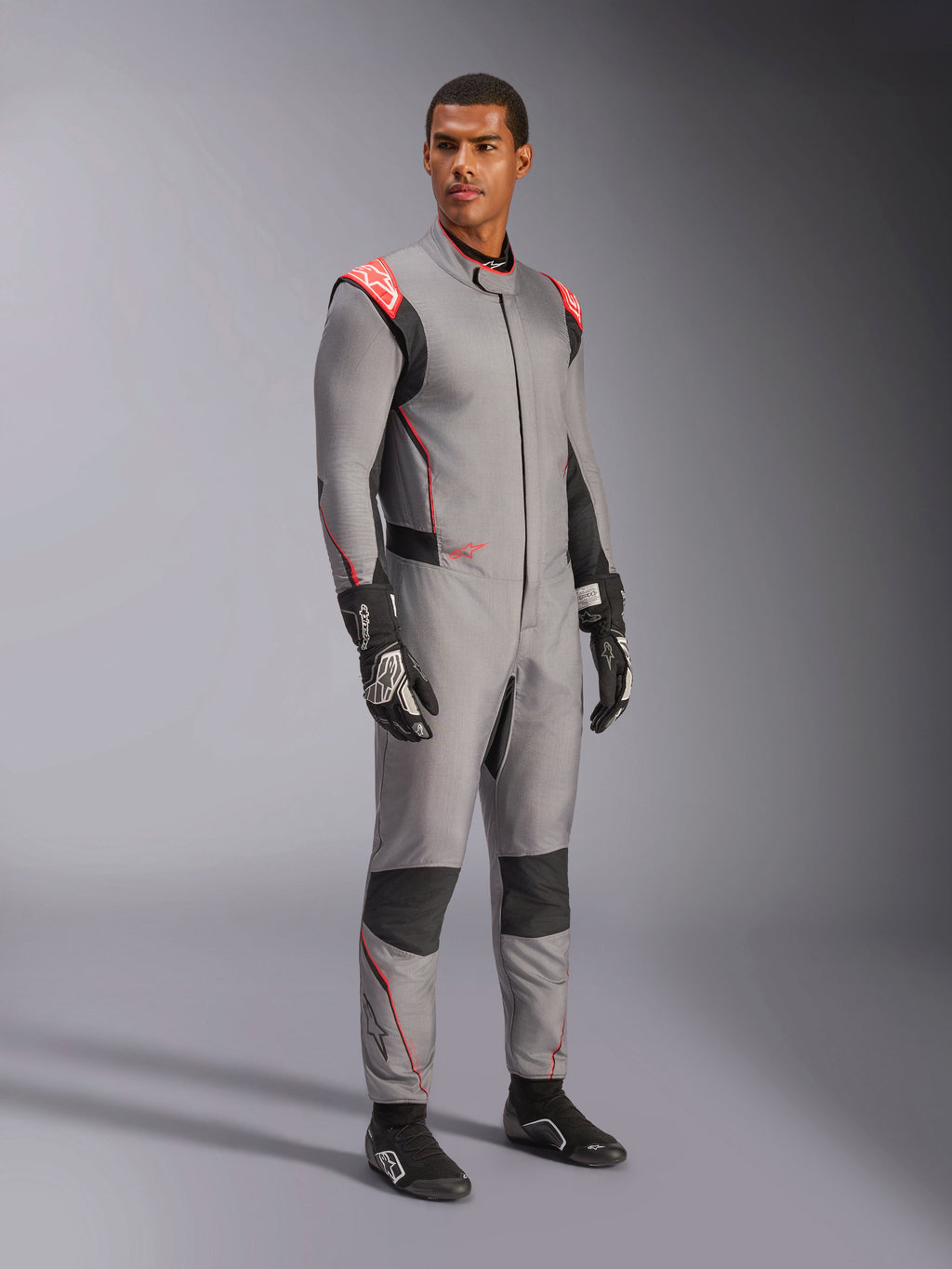 Manlig modell som bär Alpinestars Hypertech V3 Suit i Mid Gray med svarta och Red Fluo-detaljer, professionell racingoverall kombinerad med svarta racinghandskar och skor, med en lättviktskonstruktion i två lager Nomex, röda axeldetaljer och atletisk passform.