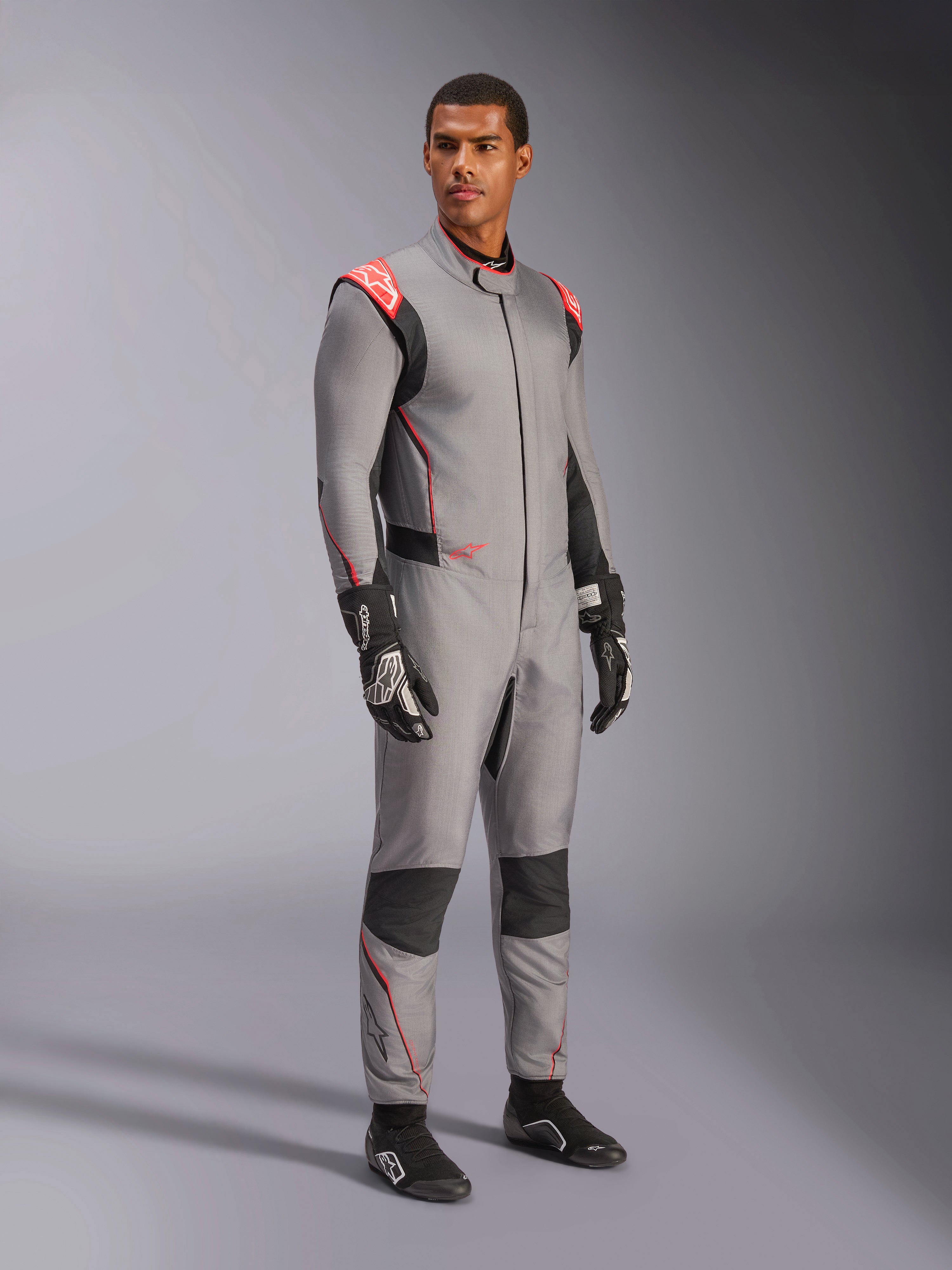 Manlig modell som bär Alpinestars Hypertech V3 Suit i Mid Gray med svarta och Red Fluo-detaljer, professionell racingoverall kombinerad med svarta racinghandskar och skor, med en lättviktskonstruktion i två lager Nomex, röda axeldetaljer och atletisk passform.