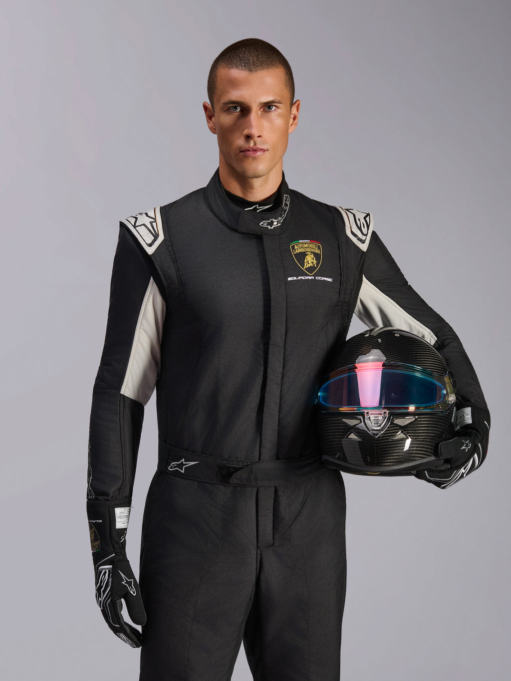 Manlig modell i Limited Edition Lamborghini GP Tech V4-dräkt, svart racingdräkt med silverfärgade detaljer och Lamborghini Squadra Corse-logotyp, håller en kolfiberhjälm, svarta racinghandskar med vita logotyper, professionell racingutrustning