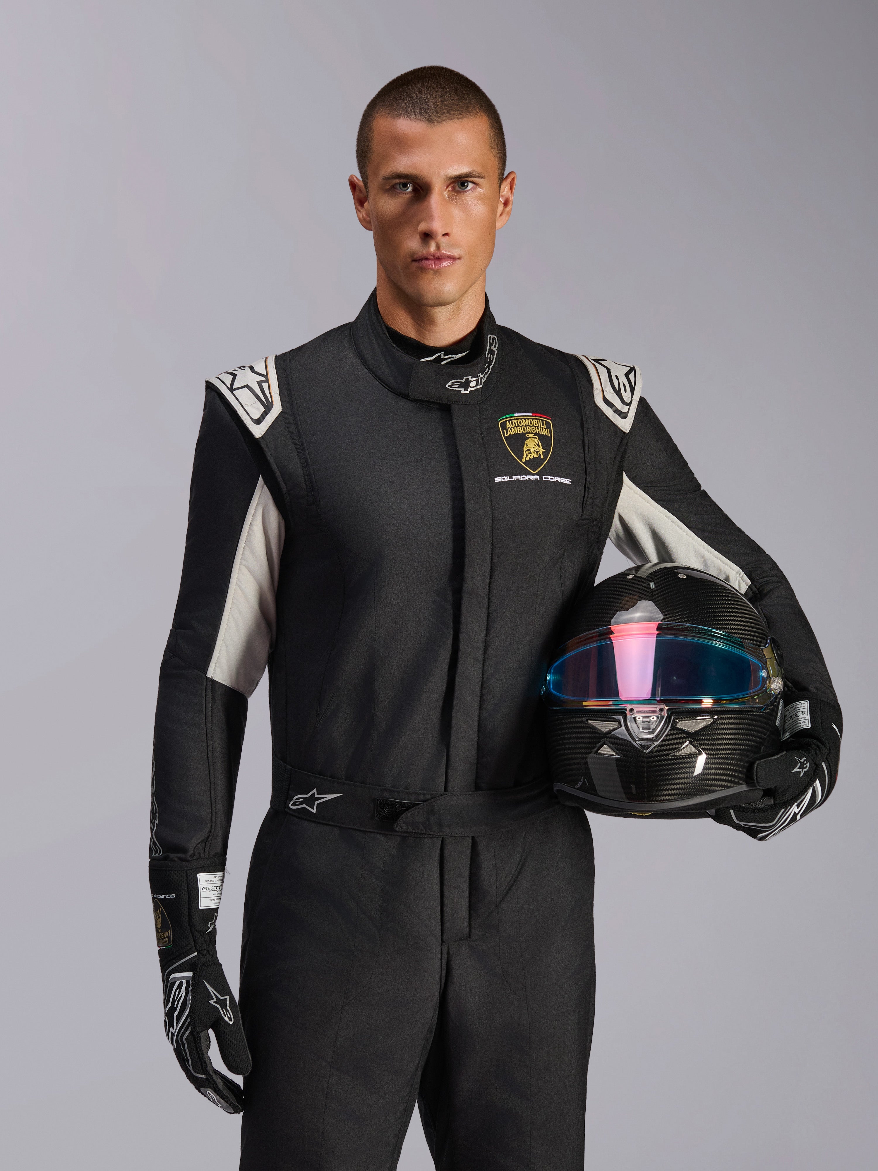 Manlig modell i Limited Edition Lamborghini GP Tech V4-dräkt, svart racingdräkt med silverfärgade detaljer och Lamborghini Squadra Corse-logotyp, håller en kolfiberhjälm, svarta racinghandskar med vita logotyper, professionell racingutrustning
