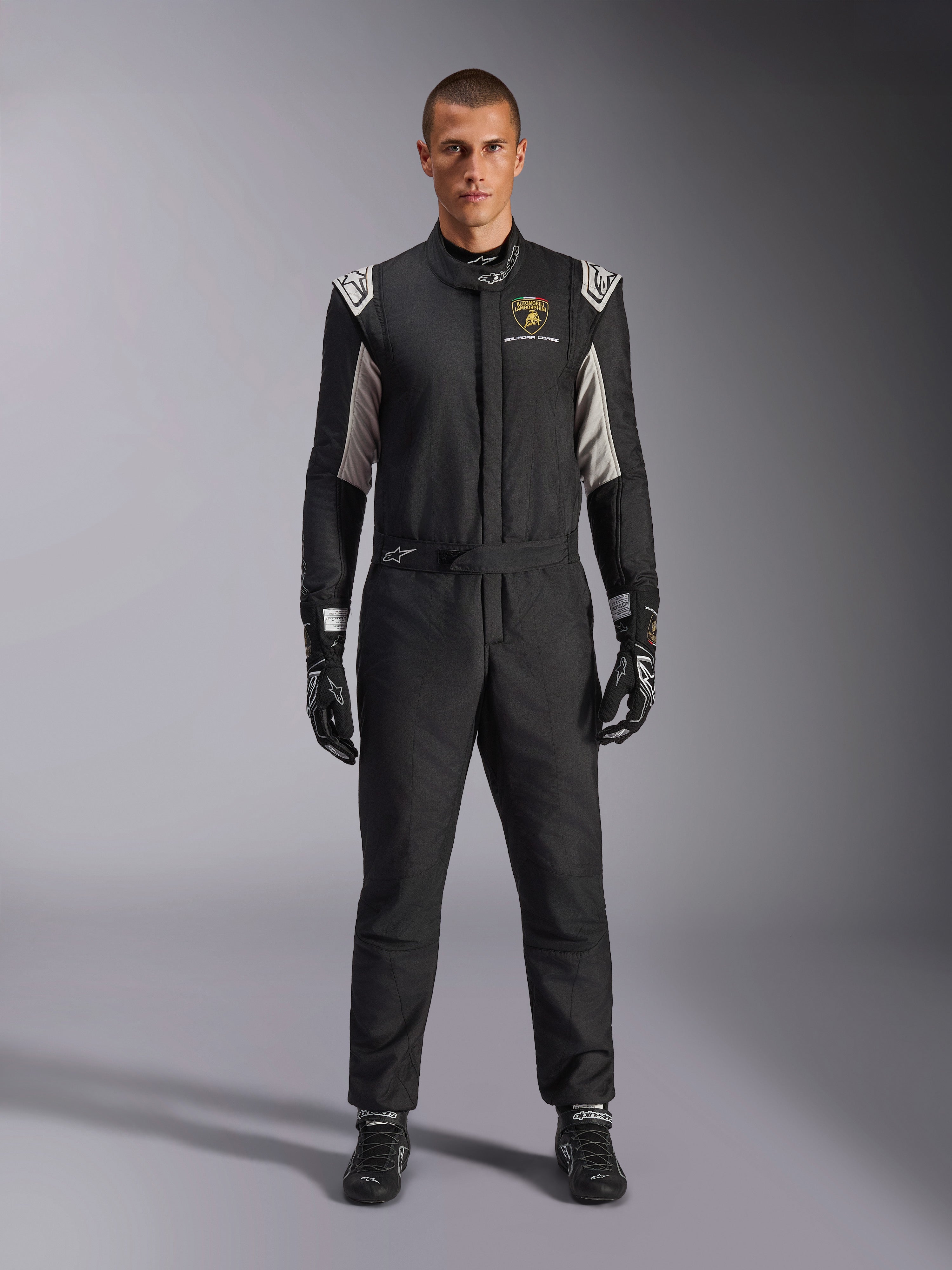 Alpinestars Limited Edition Lamborghini GP Tech V4 Dräkt FIA, professionell racingdräkt för bilsport, svart med silver- och guldfärgade detaljer, med Automobili Lamborghini Squadra Corse-logotyp på bröstet, bärs med matchande racinghandskar och stövlar, stilren atletisk design för tävlingsbruk