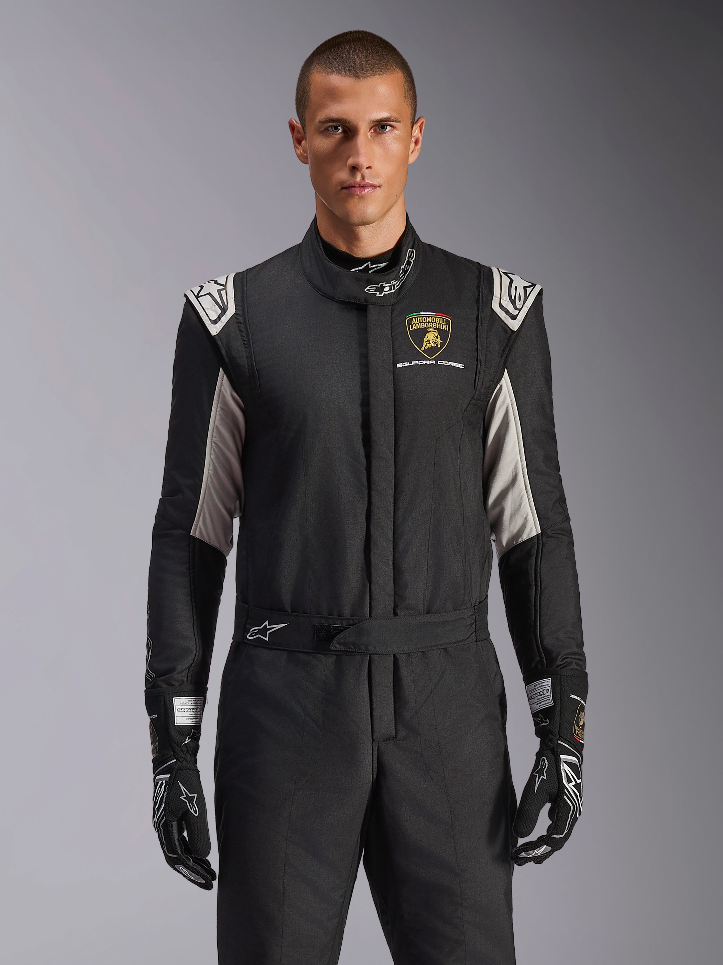 Alpinestars Limited Edition Lamborghini GP Tech V4 Suit, professionell racingdräkt i svart med silver- och gulddetaljer, med Automobili Lamborghini Squadra Corse-skölden på bröstet, bärs av en modell med matchande svarta racinghandskar.