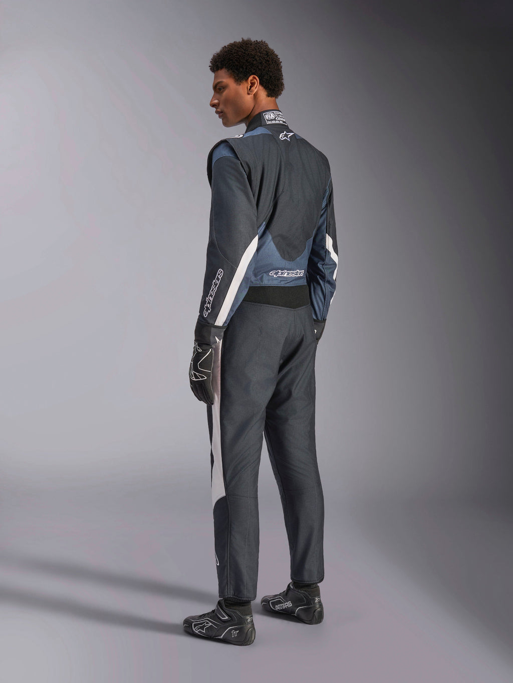 Alpinestars GP Pro Comp V2 racingoverall, auto racing för herrar, svart och asfalt med vita detaljer, FIA-certifierad, herrmodell sedd i 3/4-profil bakifrån med matchande handskar och skor, premium textilkonstruktion.