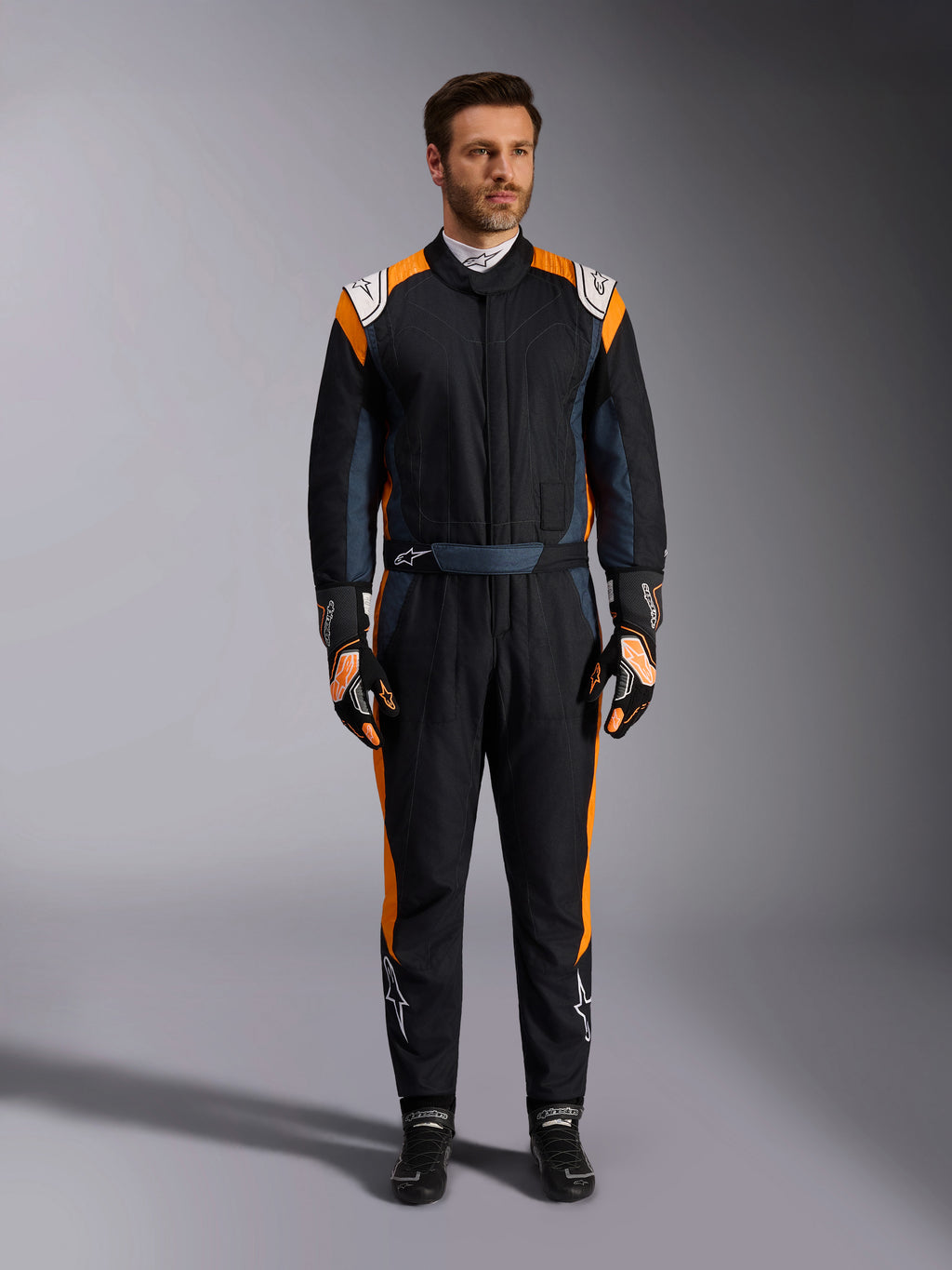 Alpinestars GP Pro Comp V2 FIA Suit, bilsportsoverall i Black Asphalt och Orange Fluo, manlig modell iklädd komplett skyddsutrustning med matchande racinghandskar och skor, vita Alpinestars-logotyper och livfulla orangea sidopaneler, professionell motorsportutrustning