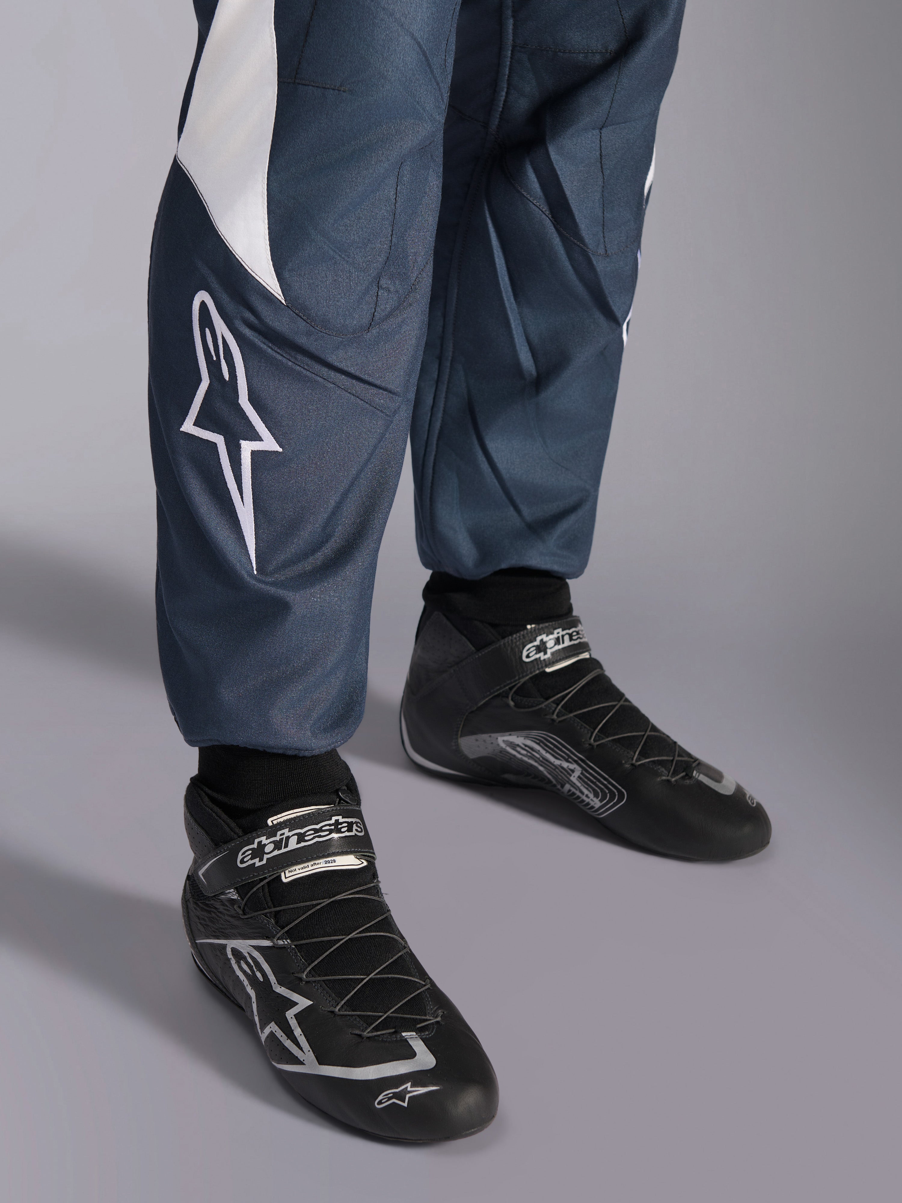 Alpinestars GP Pro Comp V2 racingoverall, FIA-godkänd bilracing, asfaltgrå och cyan, närbild på logotyp och svarta racingskor.