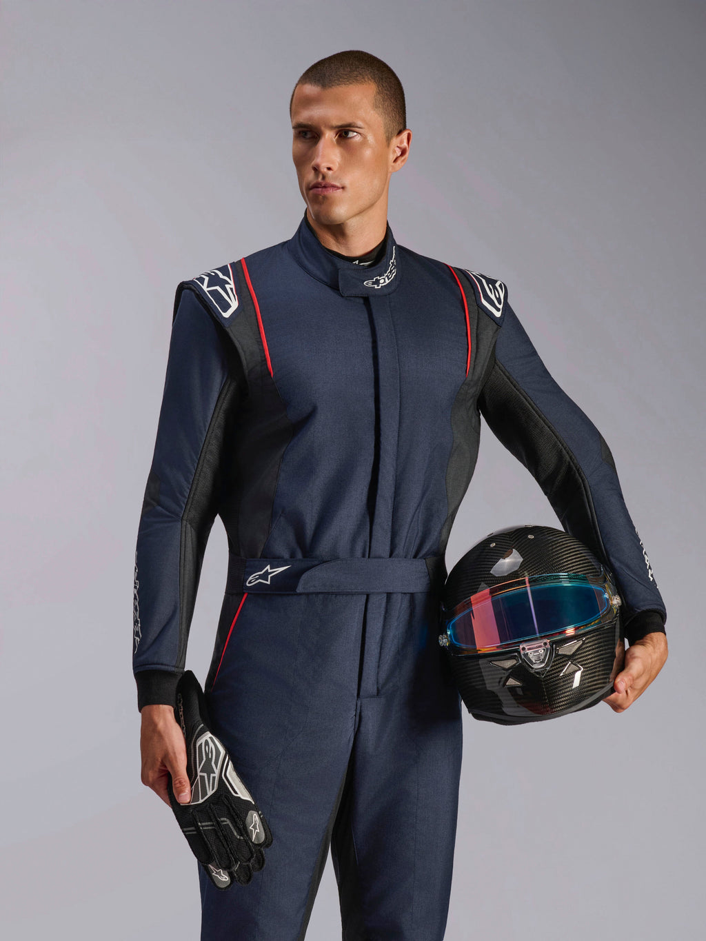 Manlig modell bär Alpinestars GP Tech V4 Suit, marinblå racingoverall med svarta och röda detaljer, håller en kolfiberhjälm och racinghandskar, professionell skyddsutrustning för motorsport