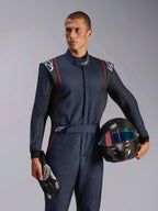 Manlig modell bär Alpinestars GP Tech V4 Suit, marinblå racingoverall med svarta och röda detaljer, håller en kolfiberhjälm och racinghandskar, professionell skyddsutrustning för motorsport