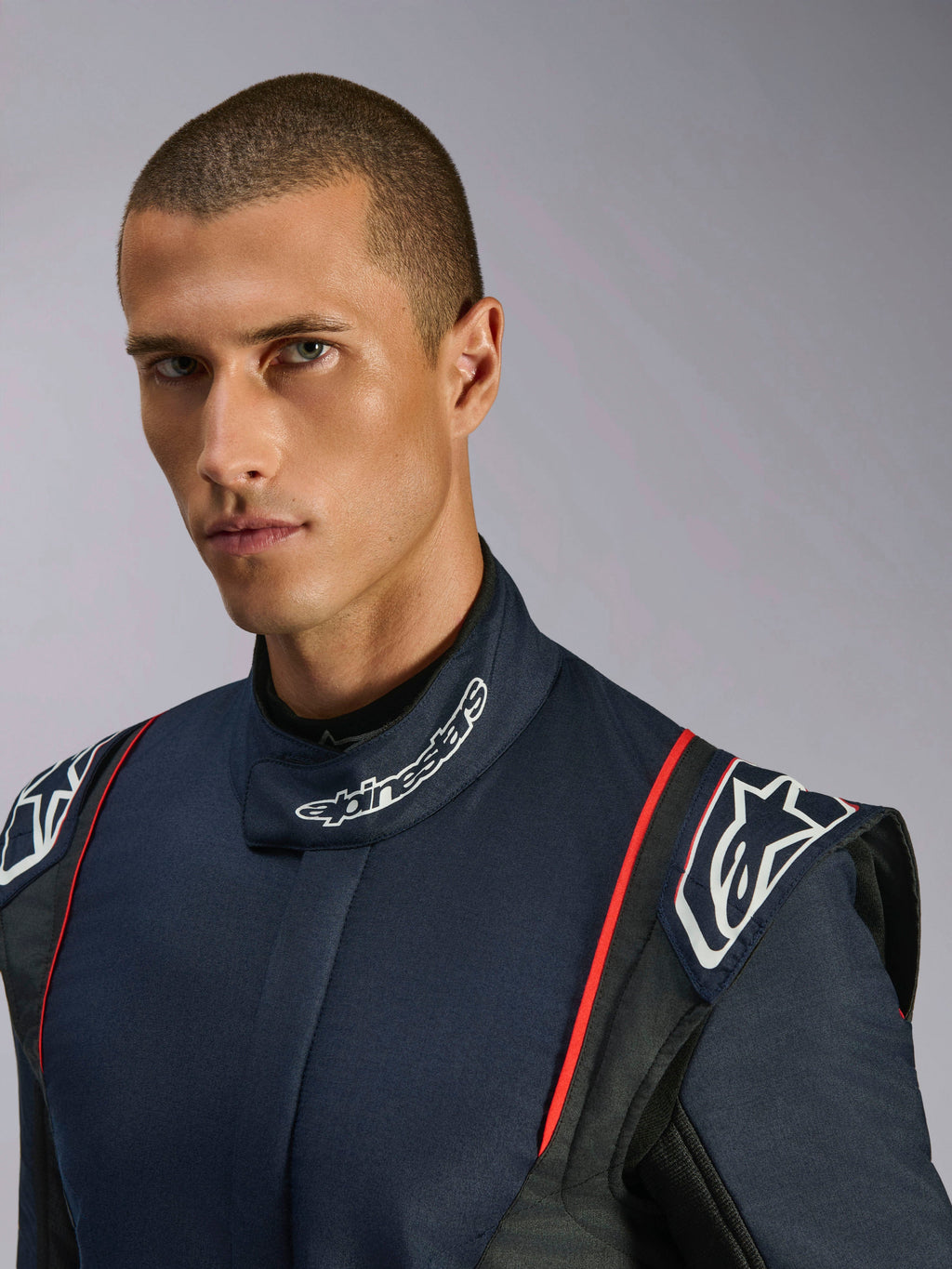 Närbildsporträtt av en modell som bär Alpinestars GP Tech V4 Suit, racingoverall för bil i mörkblått, svart och rött, med Alpinestars-märkning på den höga kragen och strukturerade axelklaffar.